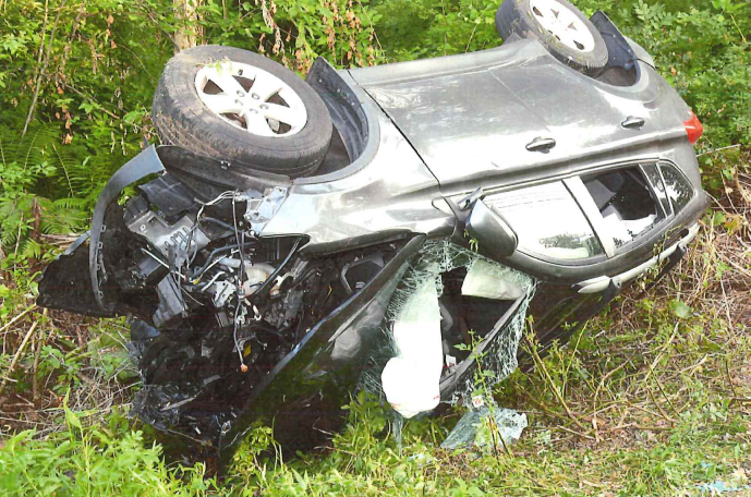 A silver car is overturned and crashed into a grassy area with trees, showing significant damage.