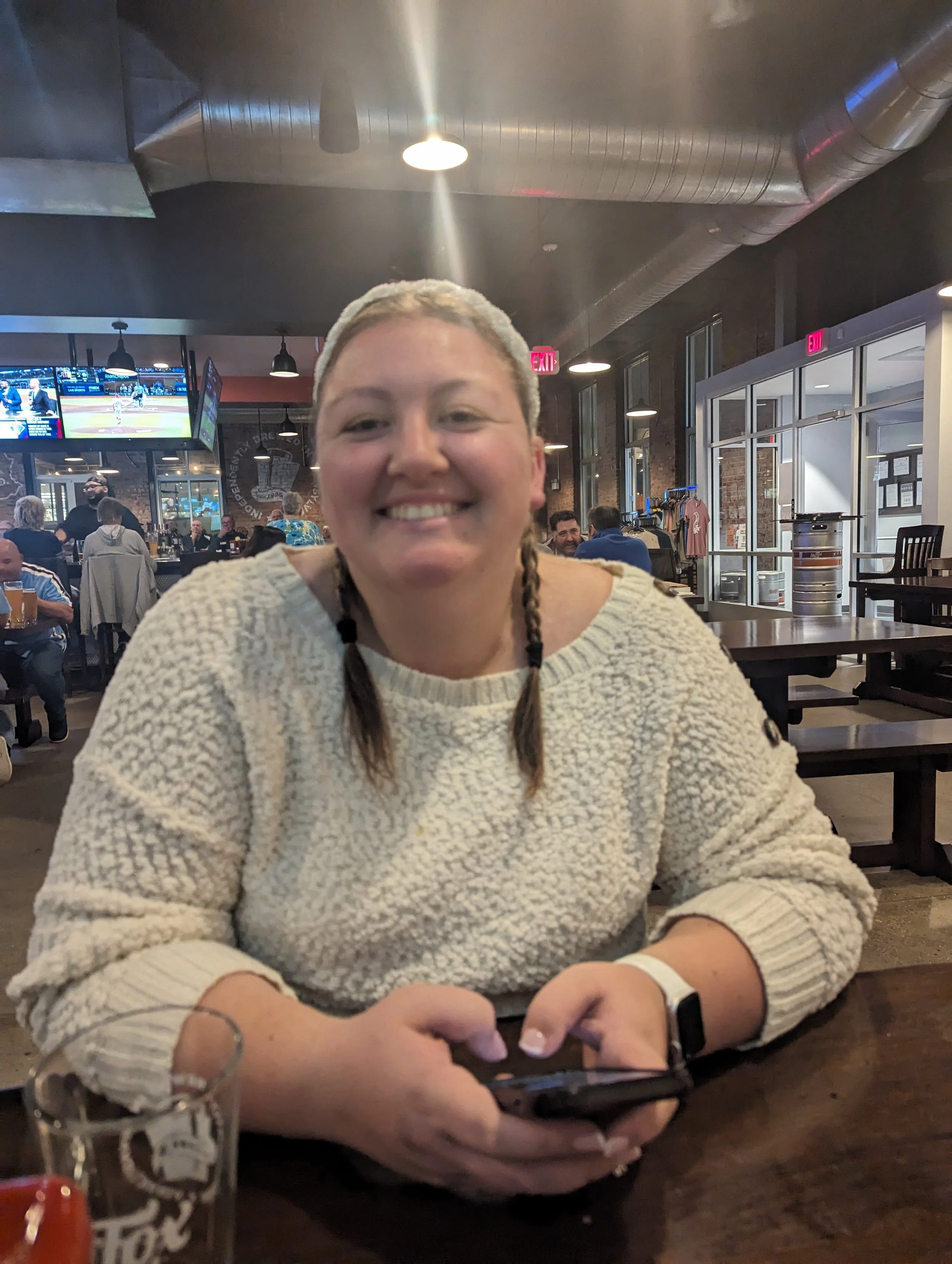 A woman with light hair in two braids smiling at the camera, sitting at a restaurant table with a phone in her hand, wearing a beige sweater, in a busy restaurant with TVs and other patrons in the background.