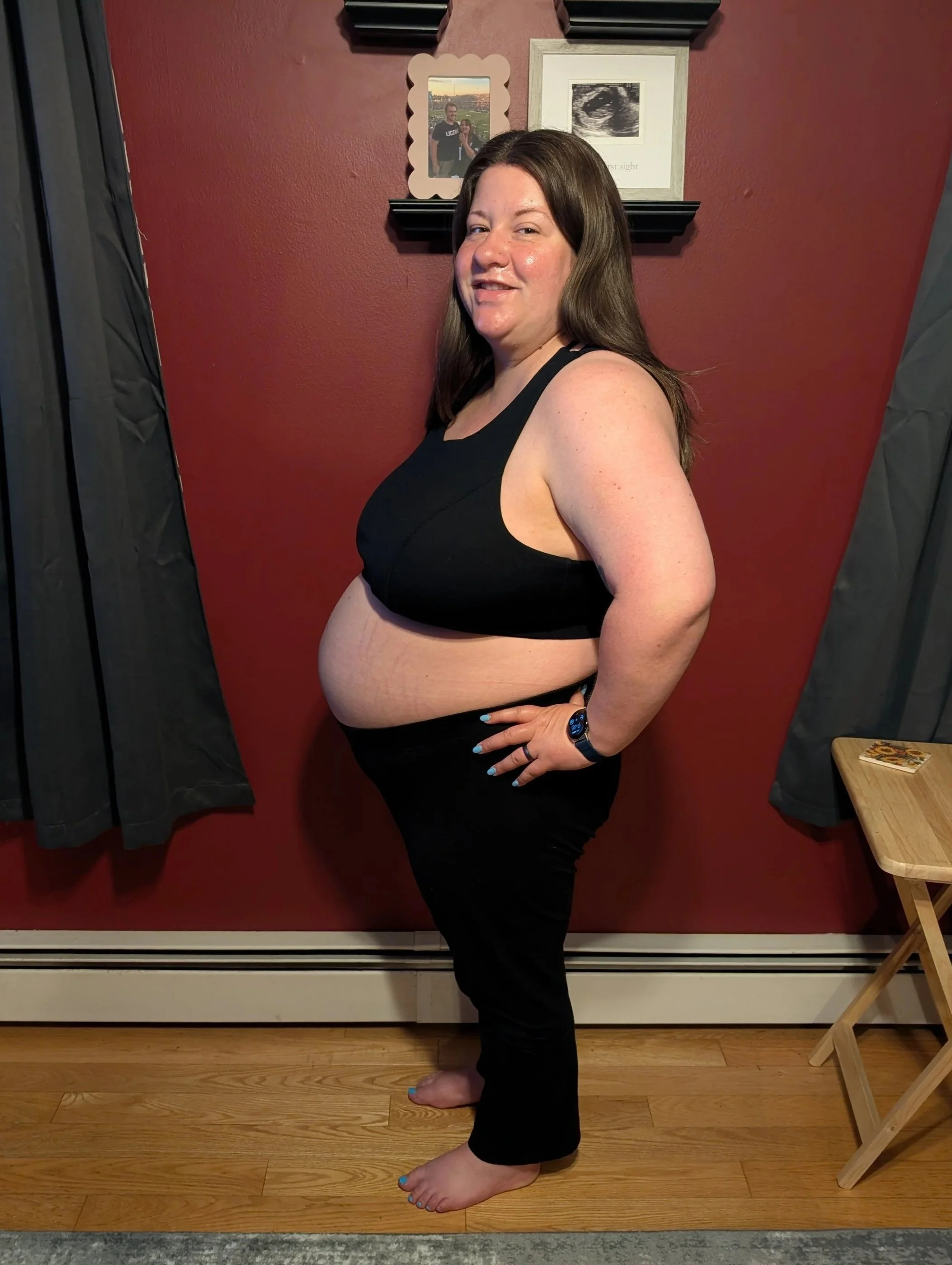 A woman standing sideways in a black sports bra and black pants, smiling, with a red wall and framed photos behind her.