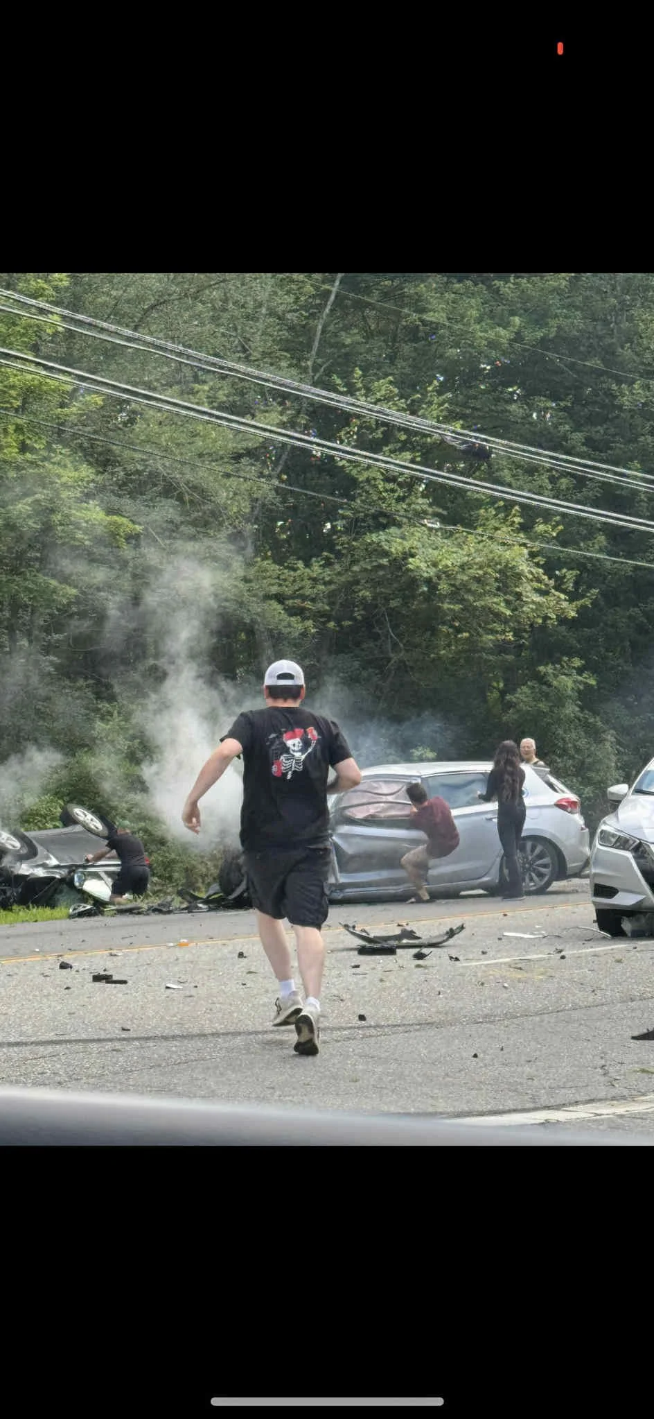 Multiple people are involved in a car accident on a road near a wooded area. Two cars are severely damaged and smoking, with debris scattered across the road. One person is kneeling near the overturned vehicle, and several others are standing near the damaged cars. A man wearing a black t-shirt with a skull and crossbones graphic and a white cap is running toward the scene.