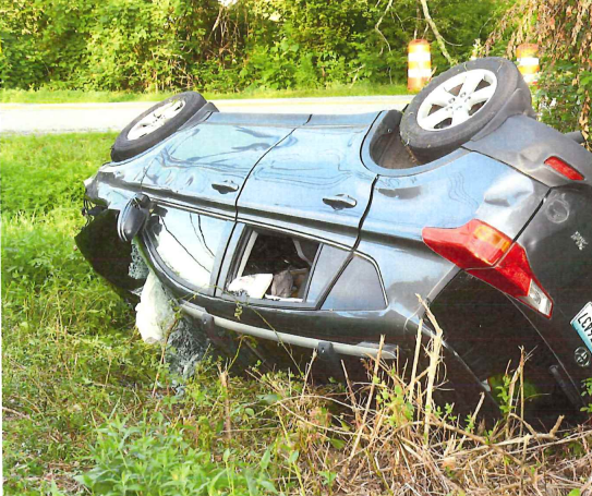 A black compact car is overturned in a grassy ditch, partially submerged, with water leaking from beneath it.