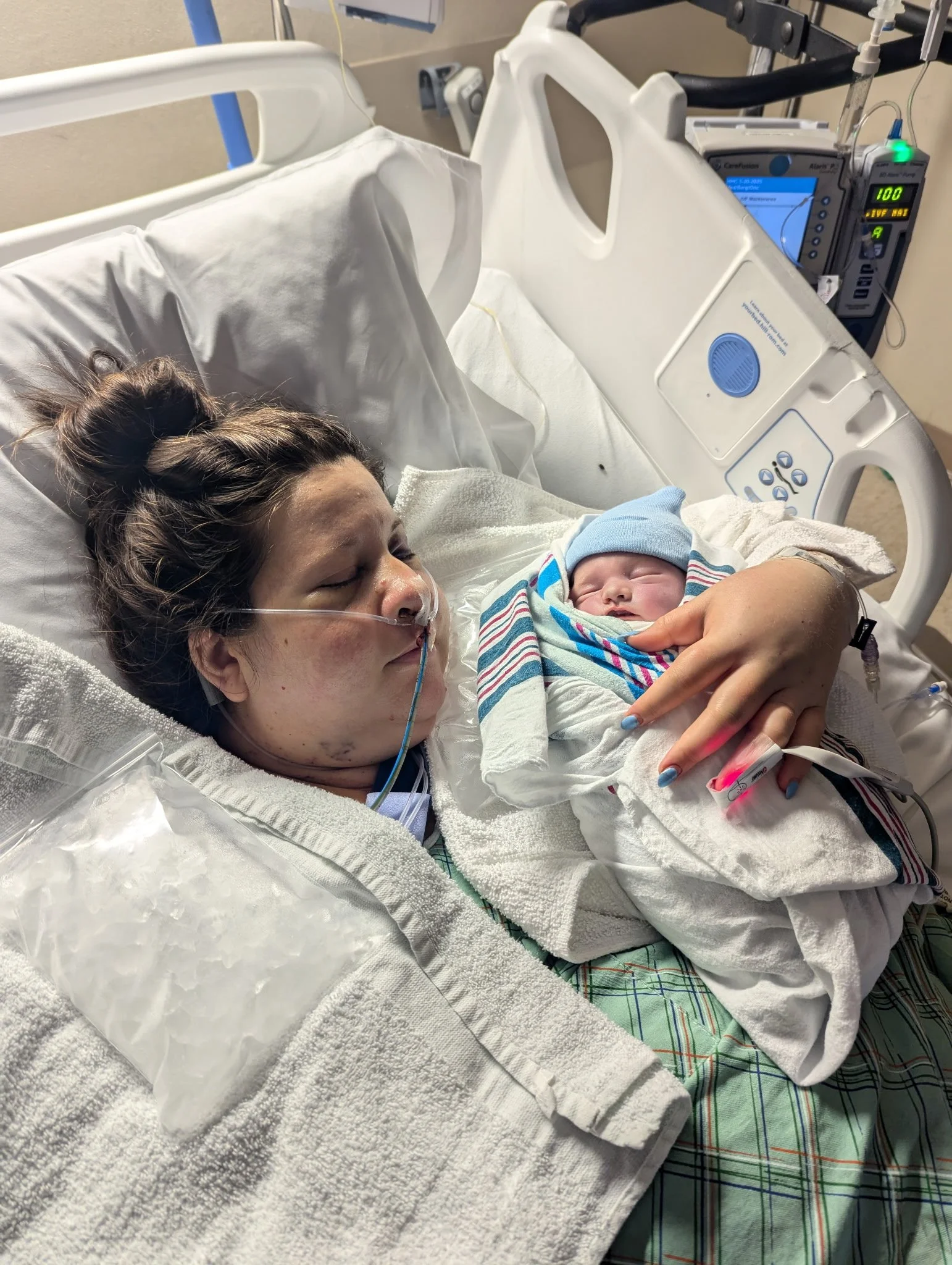 A mother lying in a hospital bed holding her newborn baby, both with eyes closed, surrounded by medical equipment.