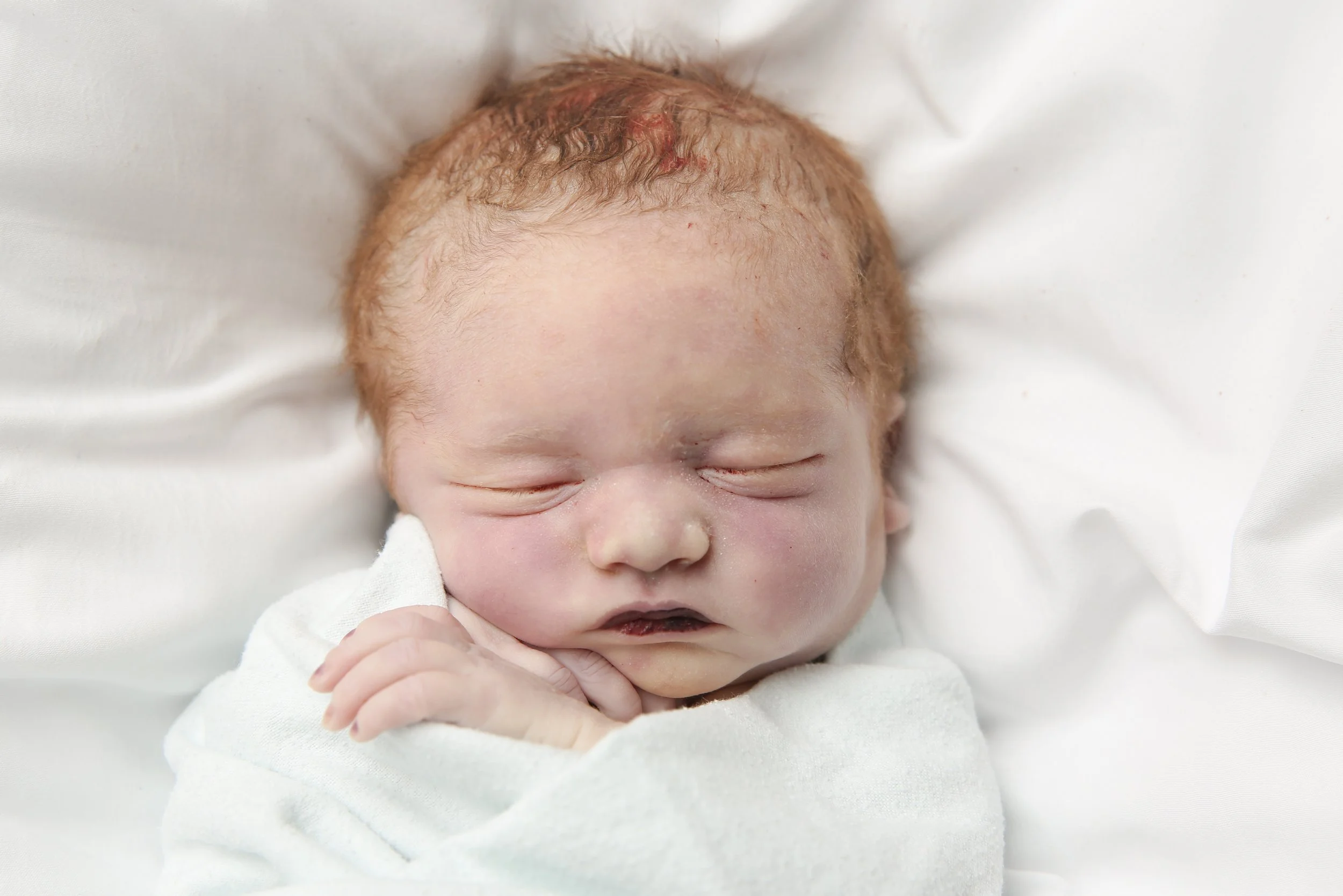 Close-up of a sleeping newborn baby with red hair, resting on white bedding, with eyes closed and hand near face.