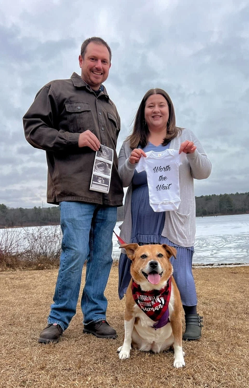 A couple standing outdoors near a lake, holding ultrasound images and a baby onesie that reads 'Worth the Wait,' with a smiling dog sitting in front wearing a bandana that says 'Parenting me is getting me all human.'
