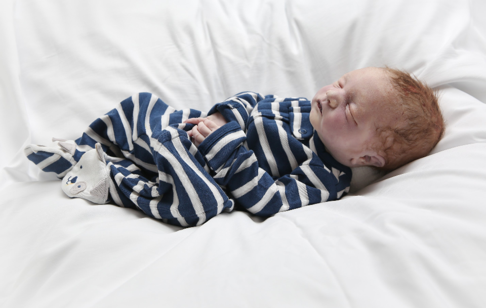 A baby with red hair sleeping on a white bed, wearing a striped blue and white onesie and sock with a bear face.