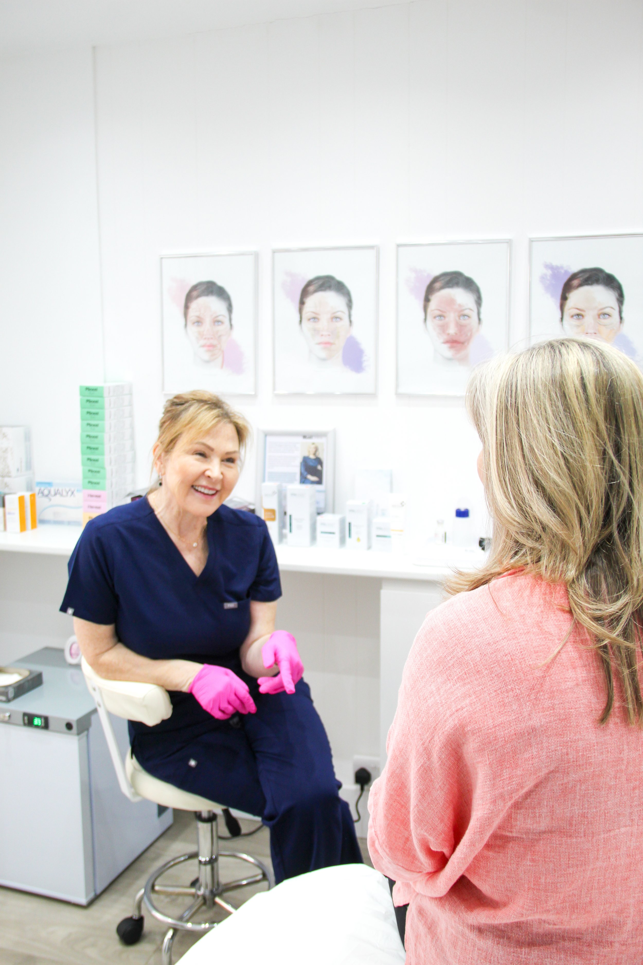 A woman wearing navy scrubs and pink gloves is sitting in a medical office, smiling and talking to another woman with long blonde hair wearing a pink sweater. The background features framed medical photographs and various medical supplies.