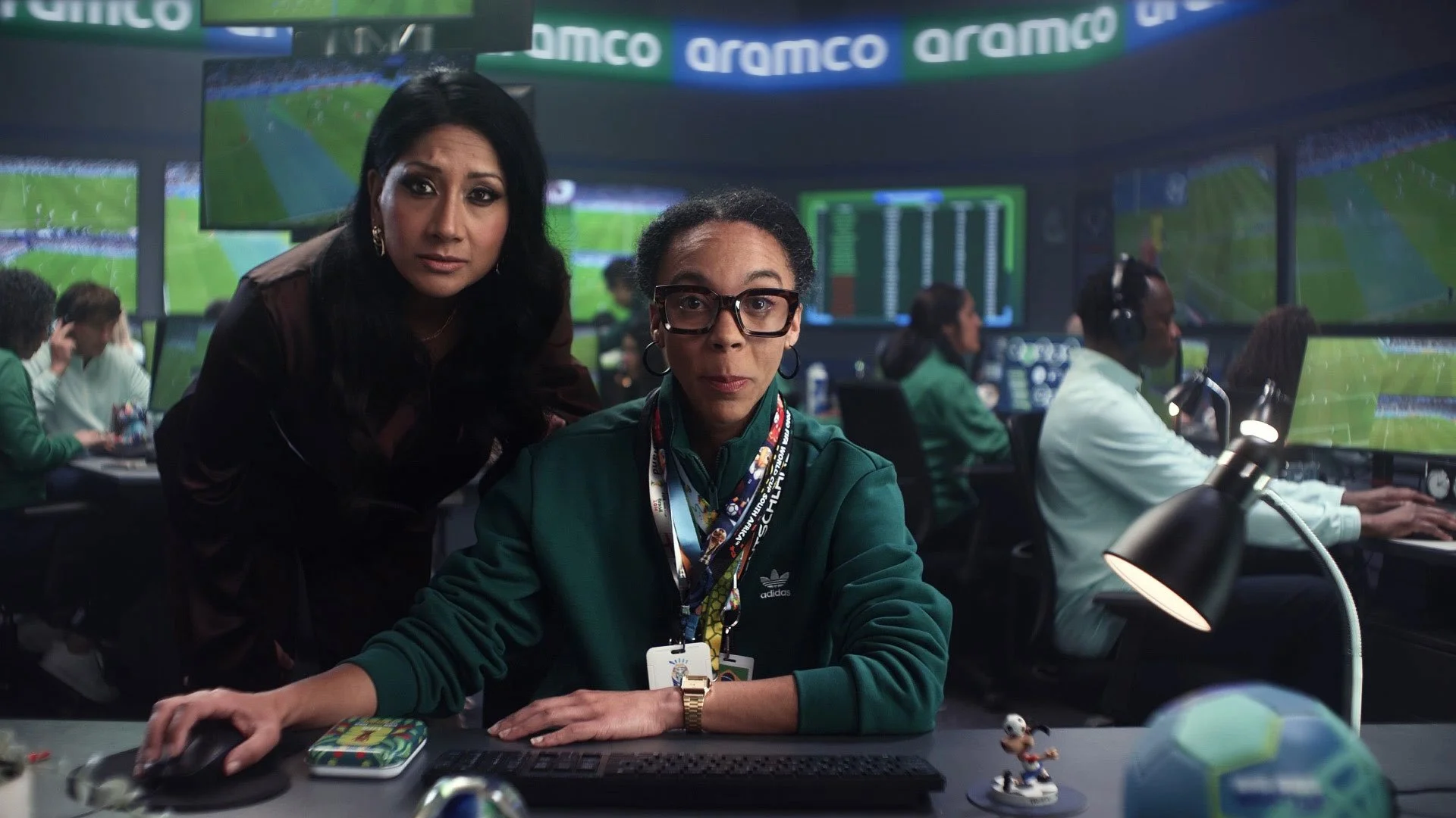 Two women in a sports control room with multiple screens displaying a football game. One woman is seated at a desk with a computer and wearing a green jacket, glasses, and an event badge; the other woman is standing beside her, leaning over. 