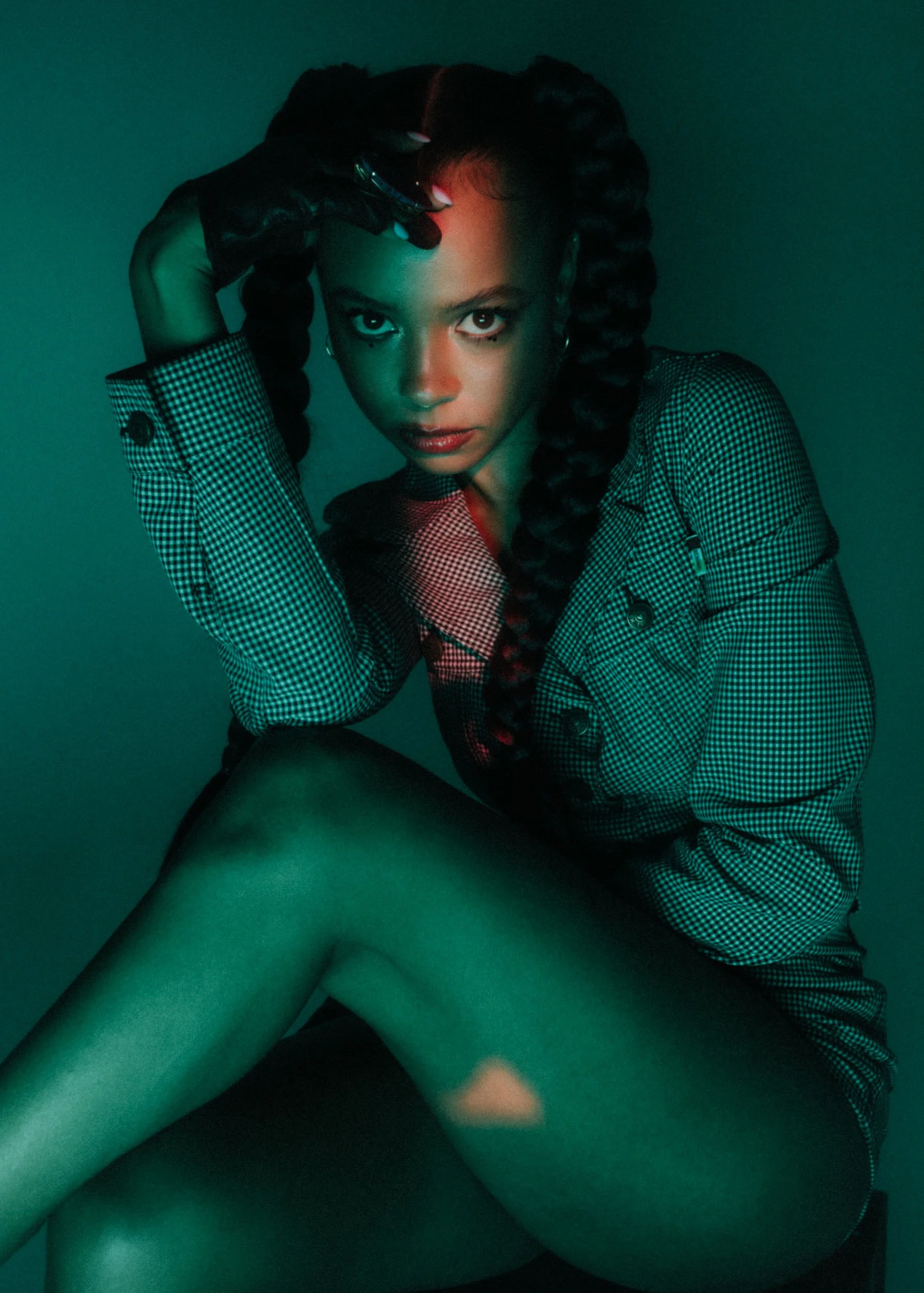 A woman with braided hair and hoop earrings poses in a dark setting with green and red lighting. She wears a checkered jacket and gloved hand touching her forehead, with her legs crossed in a provocative pose.