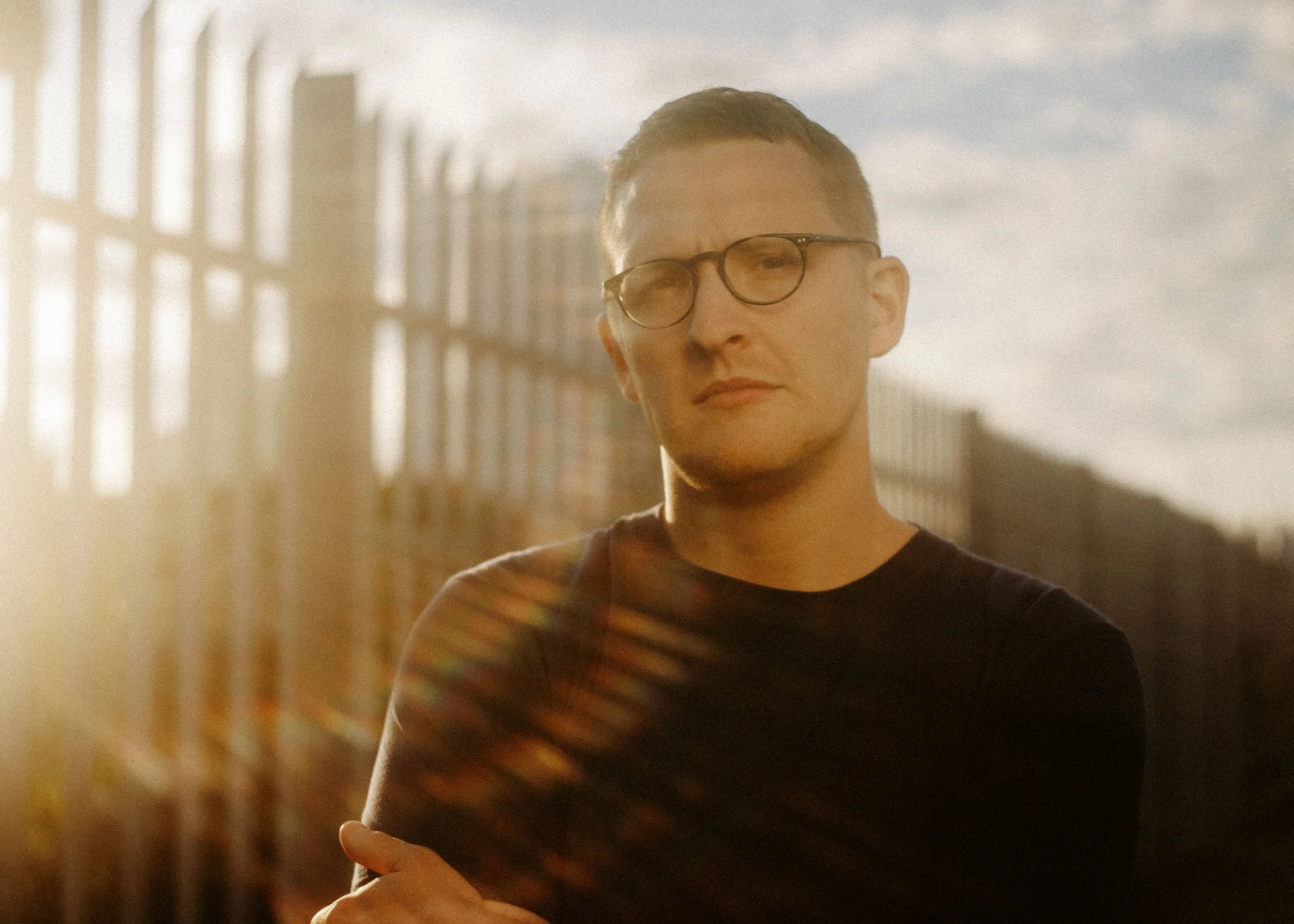A person wearing glasses with short hair, standing outdoors near a fence with a cloudy sky in the background.