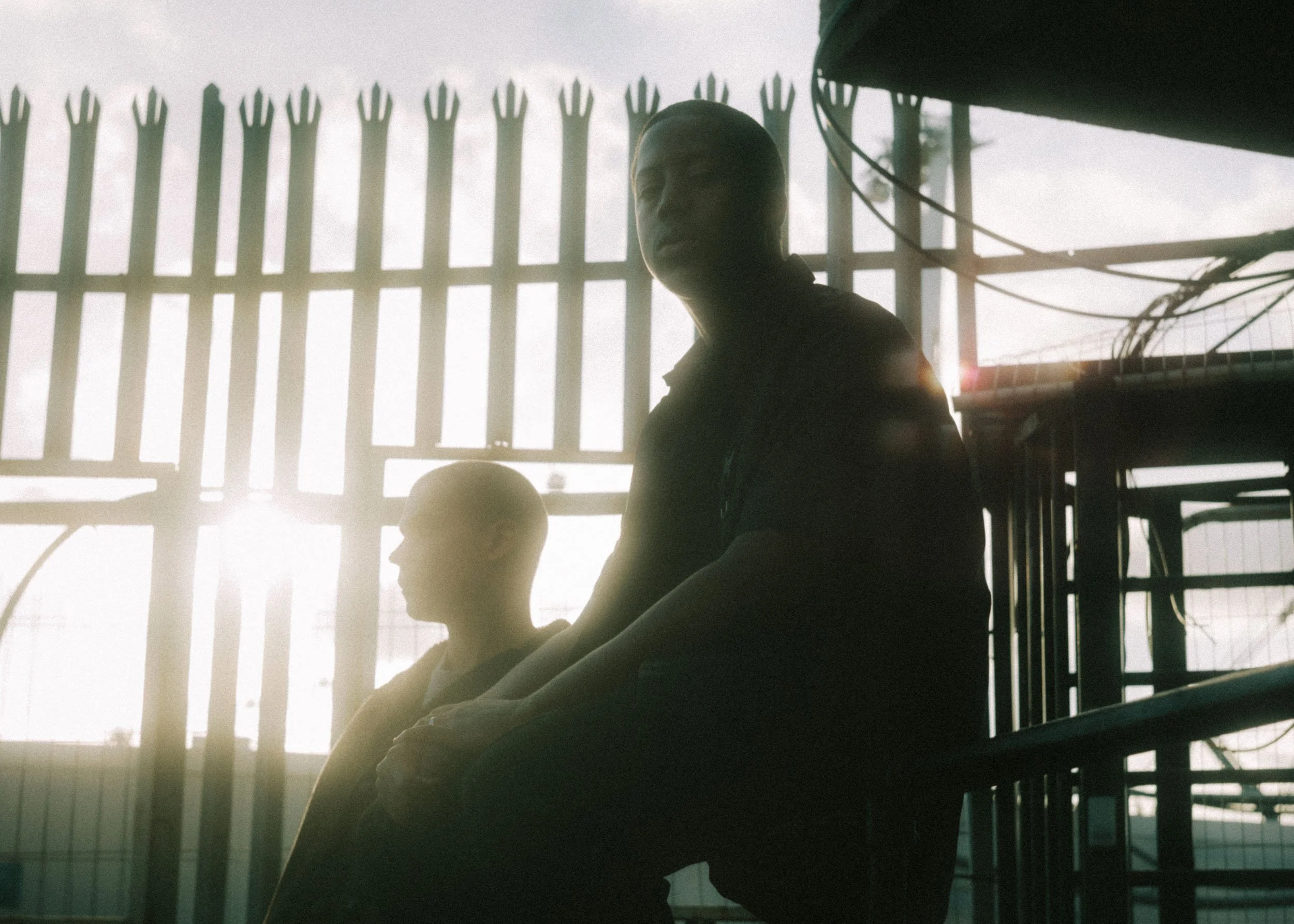 Two individuals sitting near a barred window with sunlight in the background, one appears to be a young woman and the other a young boy.