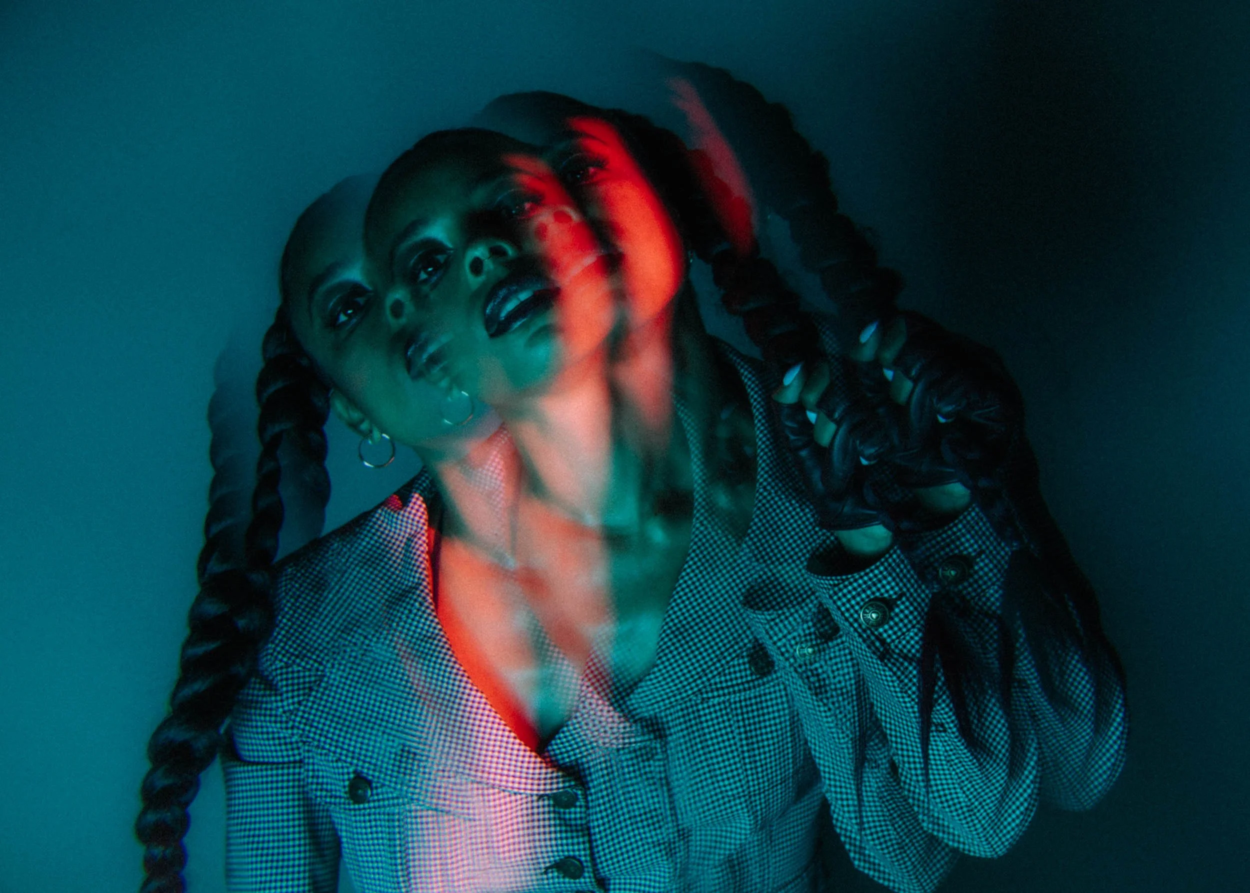 A woman with braided hair wearing a checkered blazer and turtleneck, looking up with a surprised expression. The image has a dual exposure effect with red and green lighting.
