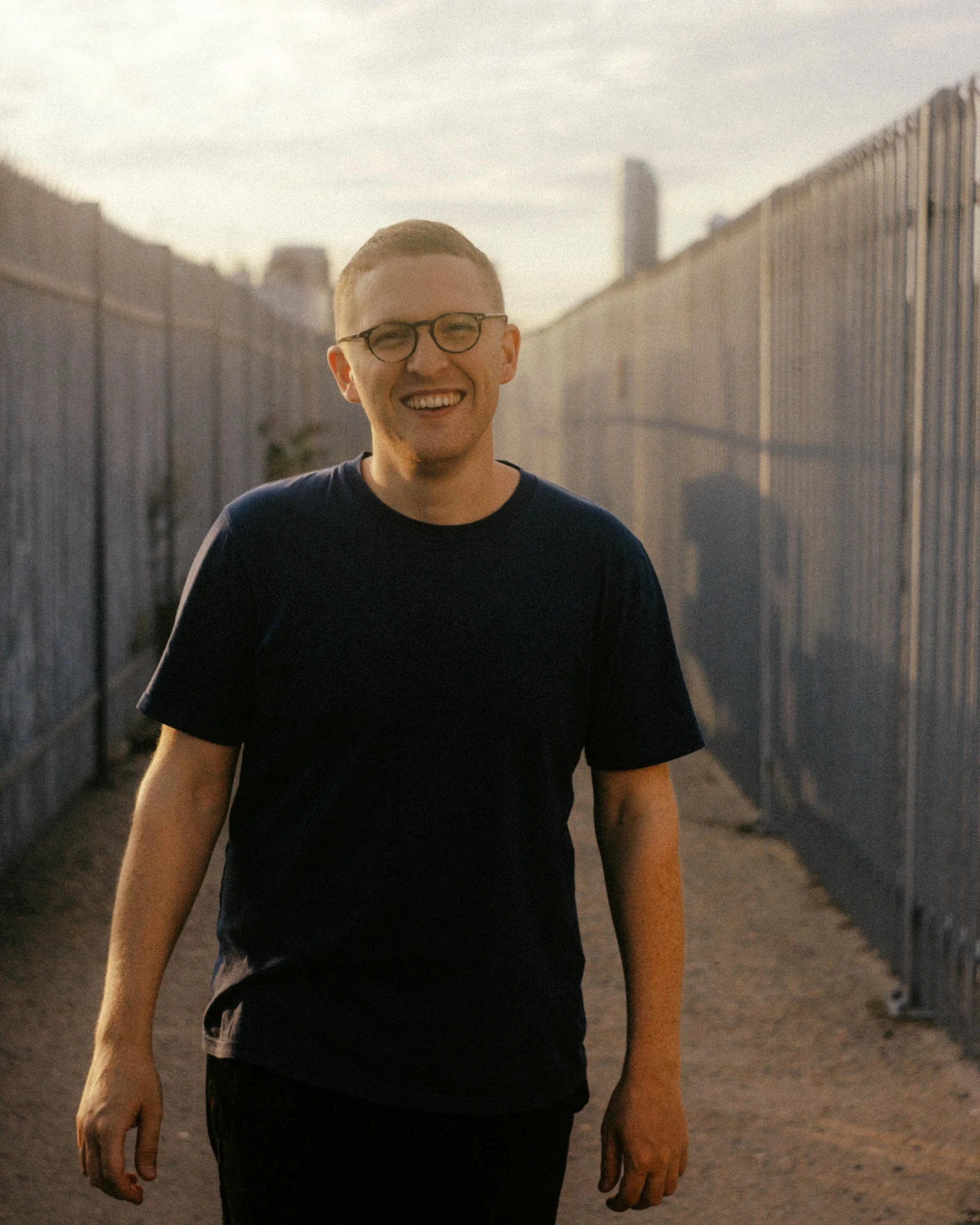 A smiling young man with glasses wearing a black T-shirt, walking outdoors between two tall chain-link fences, with city buildings in the background during sunset.