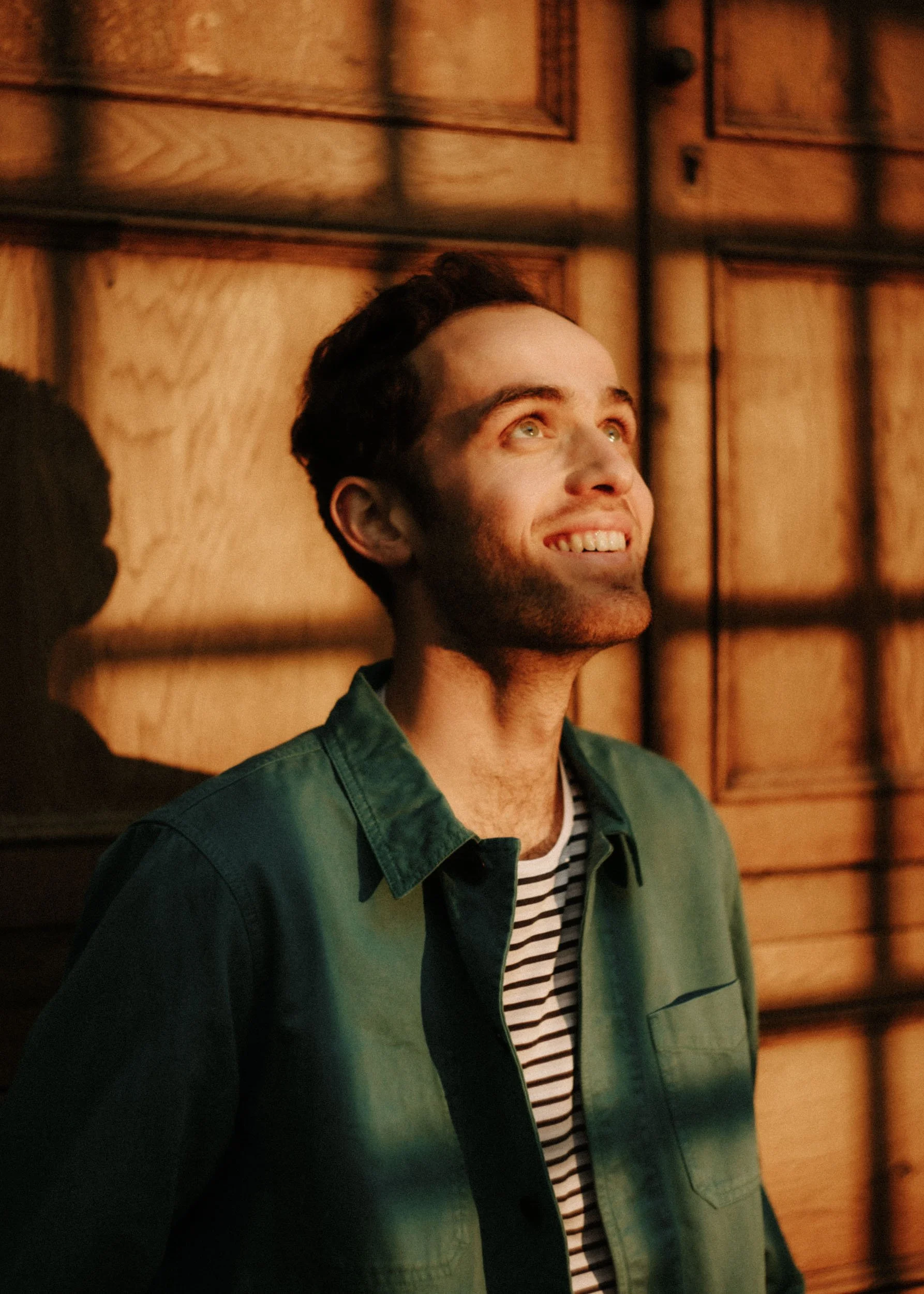 A young man with short dark hair, blue eyes, and light skin, smiling and looking up, standing in front of wooden paneling.