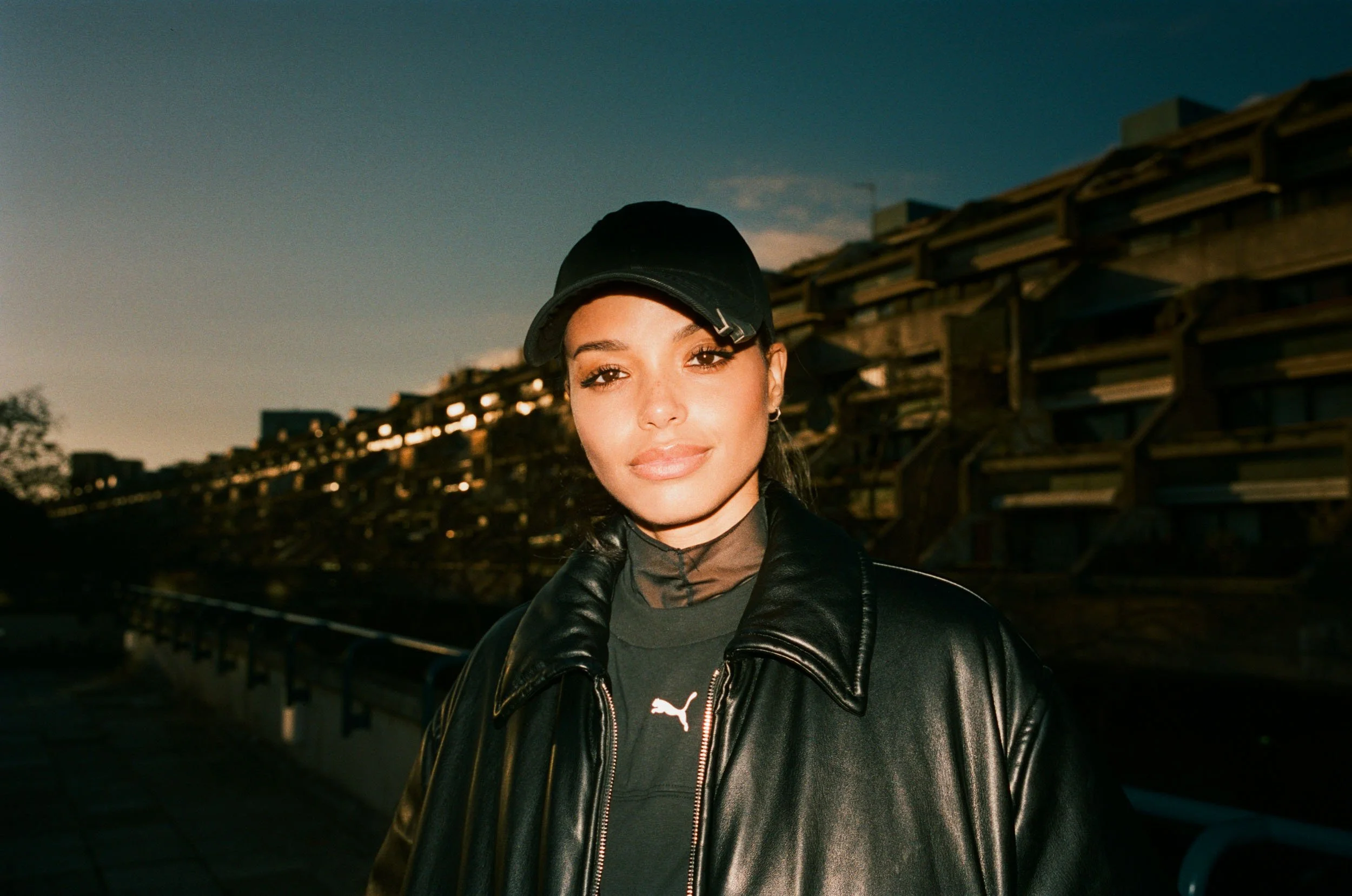 Linska with dark hair wearing a black cap and leather jacket, standing outdoors during sunset, with a structure in the background.
