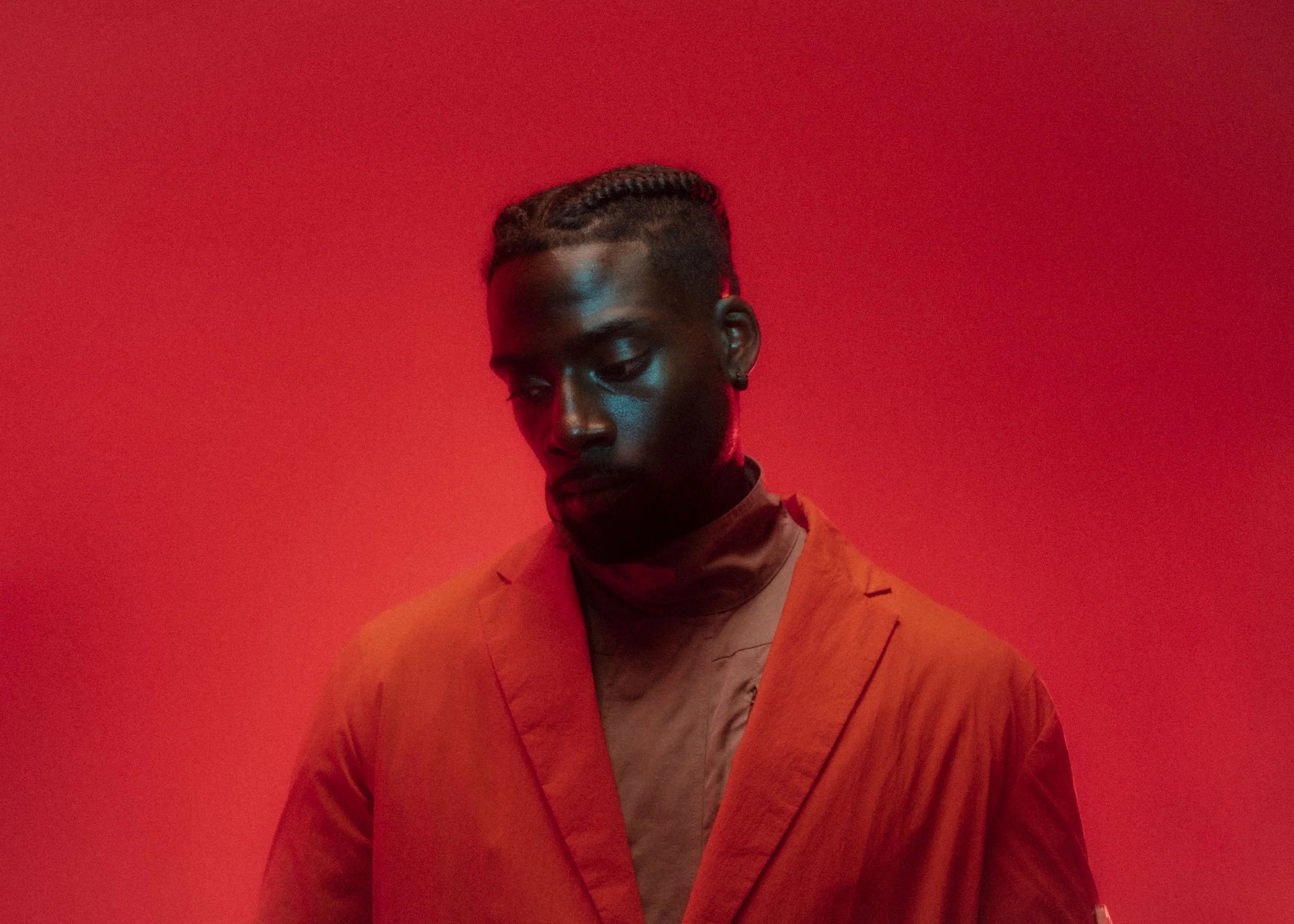 Portrait of a young man with braided hair, wearing an orange blazer and beige shirt, against a solid red background.