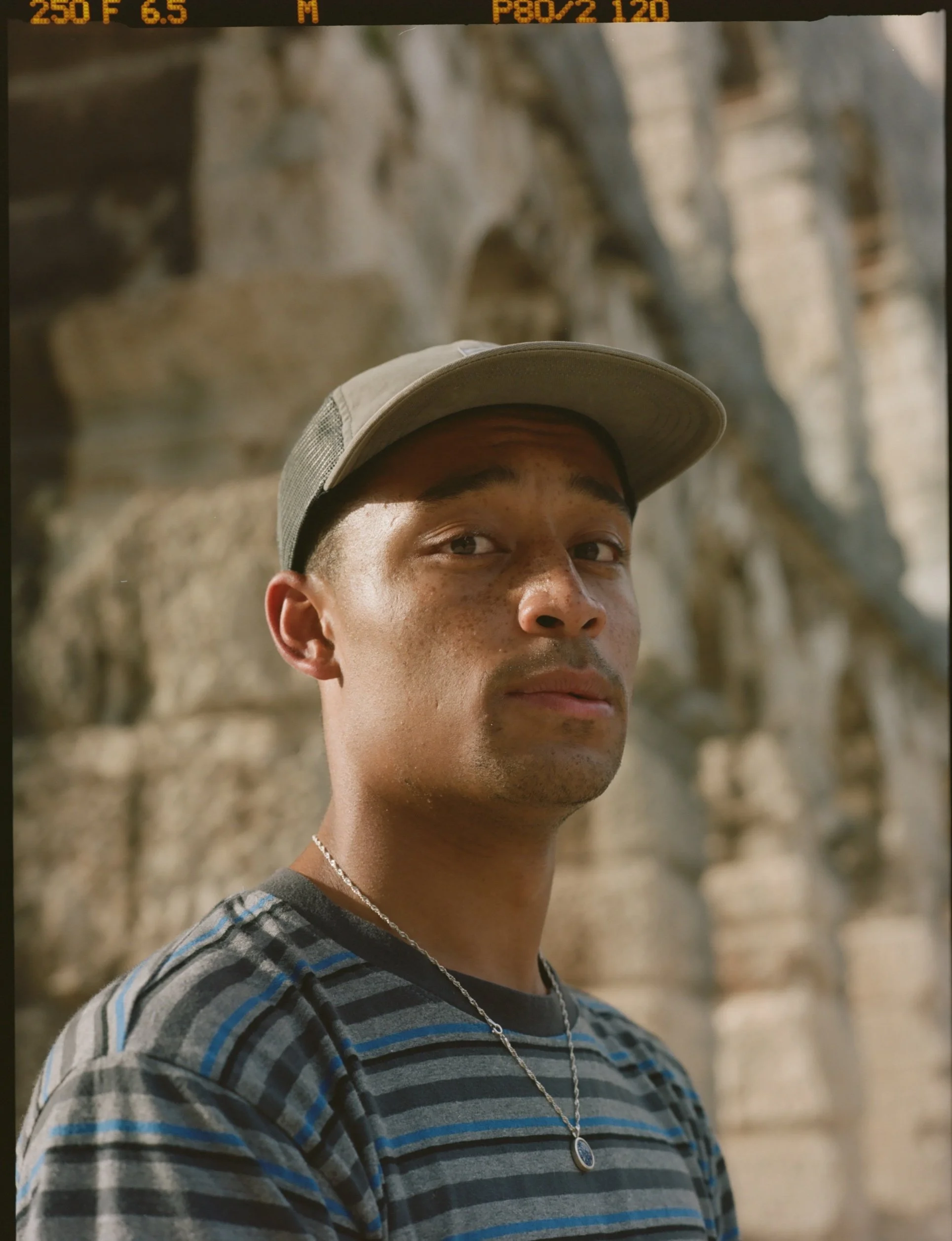 A close-up of a young man wearing a beige baseball cap, a silver necklace, and a gray and blue striped shirt. He is outdoors against a rocky background.