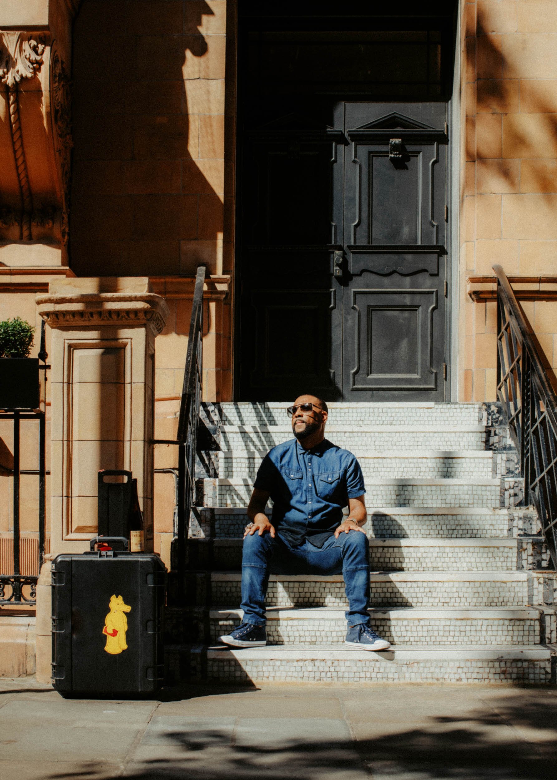 A man in a blue shirt and sunglasses sitting on the stairs in front of a black door with ornate woodwork, next to a black suitcase with a yellow teddy bear sticker, on a city sidewalk.