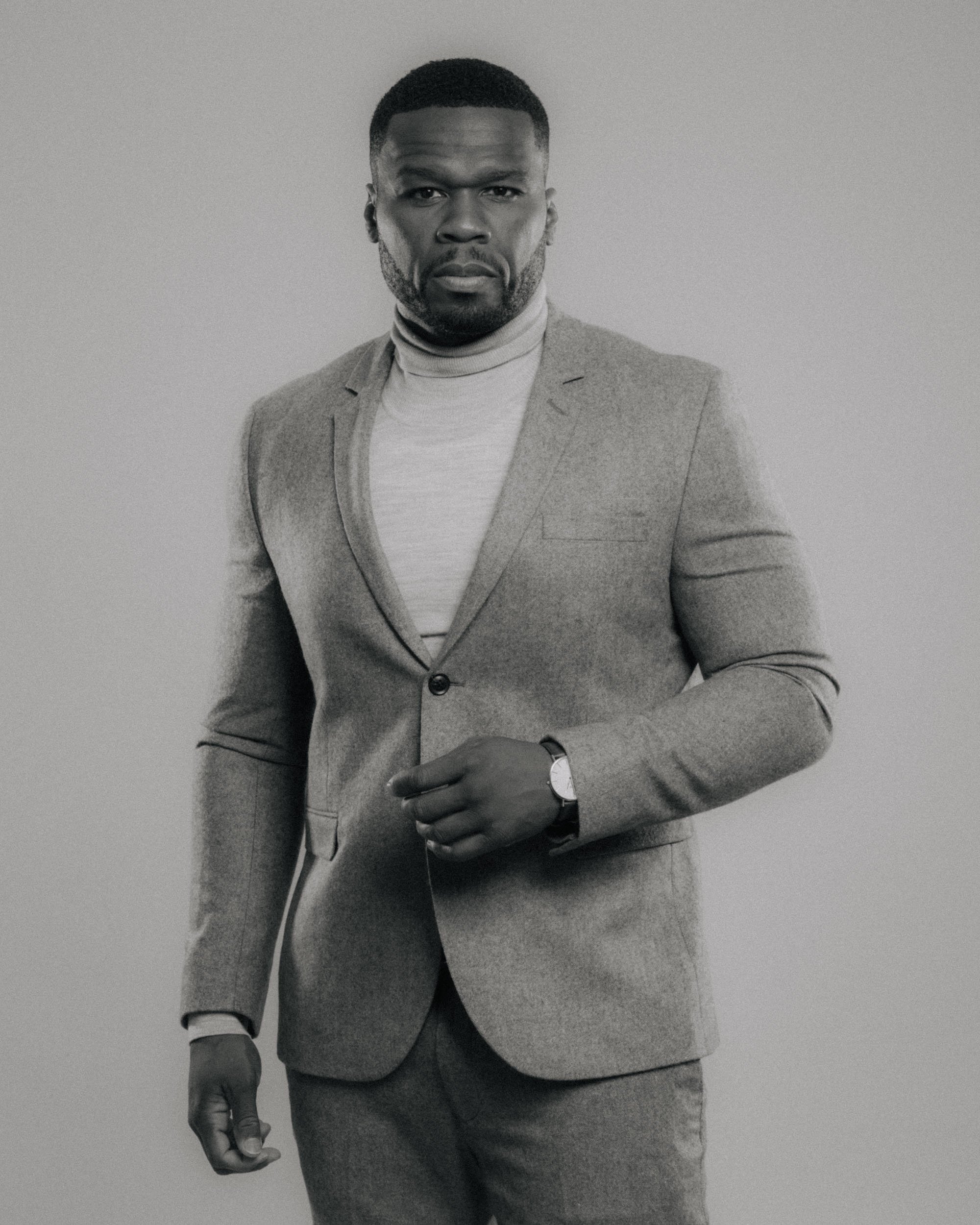 Black and white portrait of a man wearing a blazer and a turtleneck, looking serious, with his left hand gripping his right wrist.