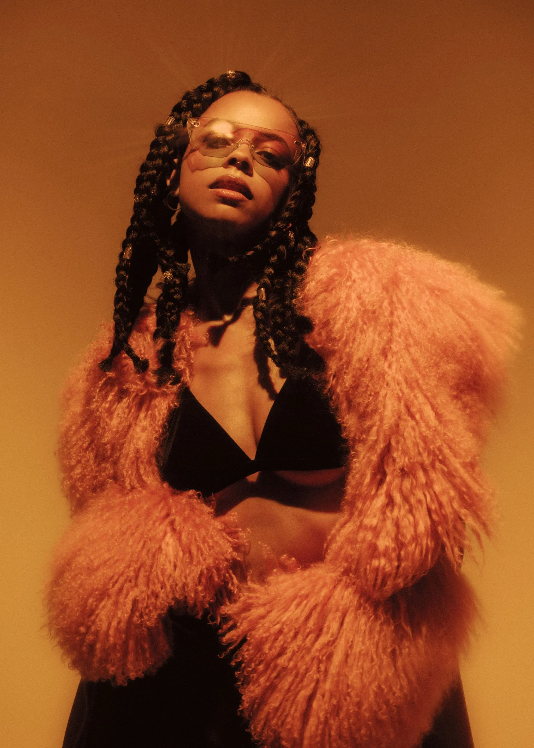 A person with dark, braided hair wearing large, tinted sunglasses, a textured pink fuzzy coat, and a black top, standing against a plain brown background.