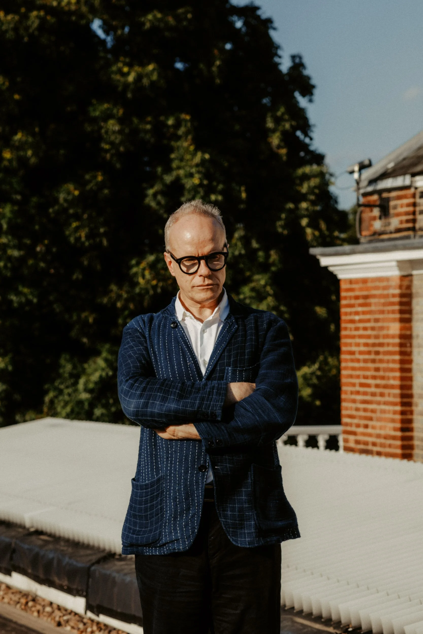 A man with glasses, light skin, and short hair wears a white shirt and a dark blue plaid blazer with his arms crossed, standing outdoors with trees and a brick chimney in the background.