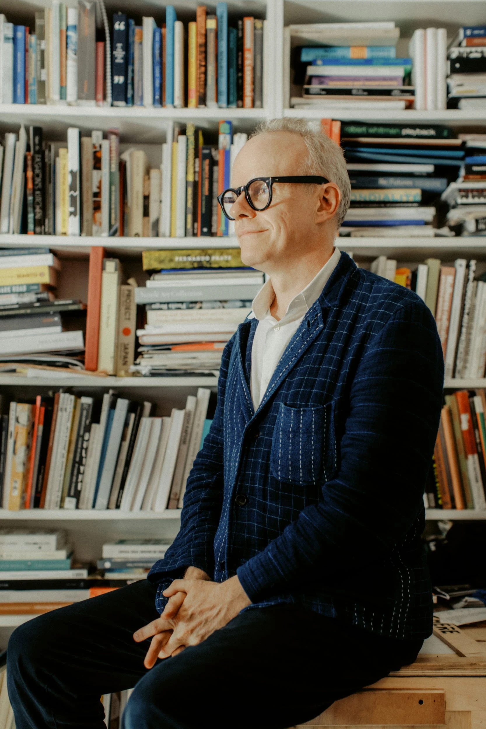 An older man wearing glasses, a white shirt, and a checkered blazer sitting in front of a bookshelf filled with books.