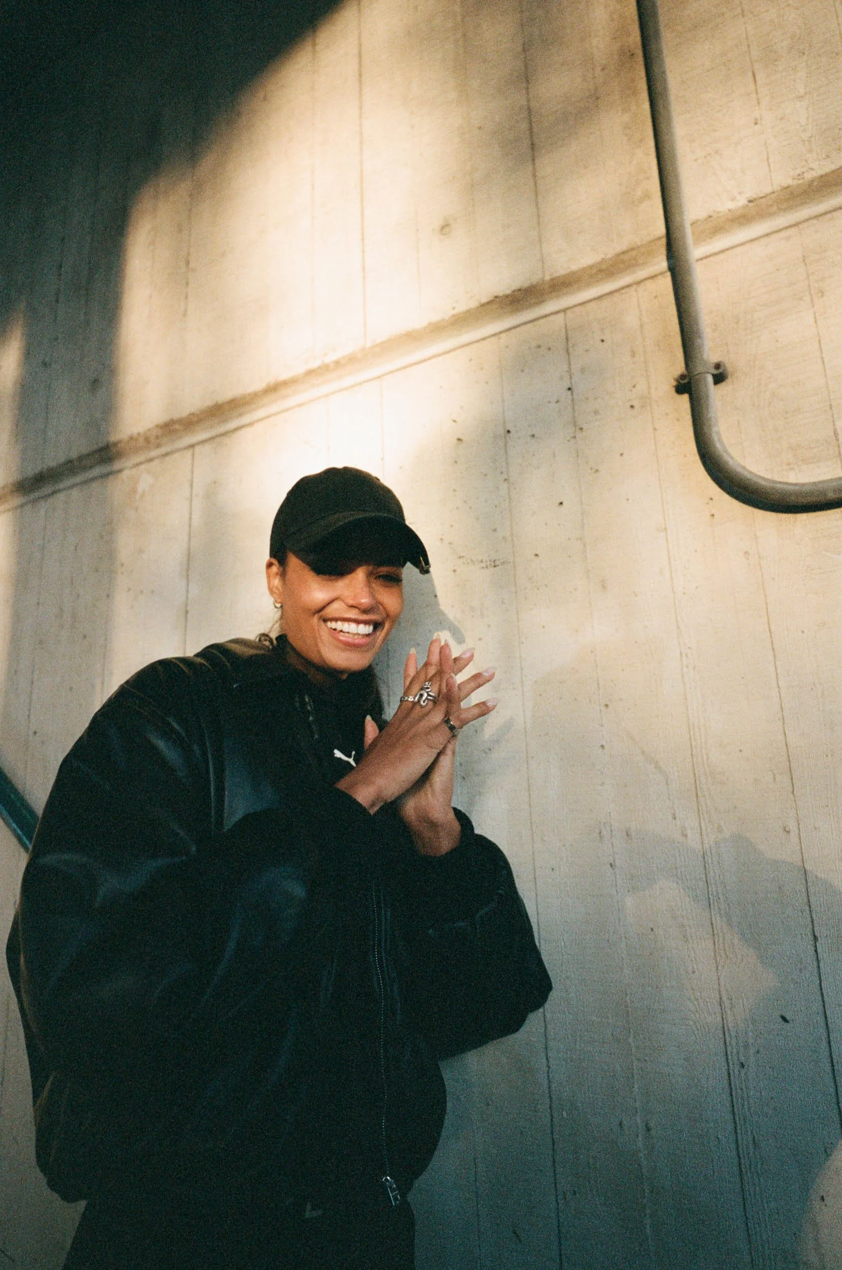 Linska in a black cap and black jacket smiling and clapping against a concrete wall with a conduit pipe.