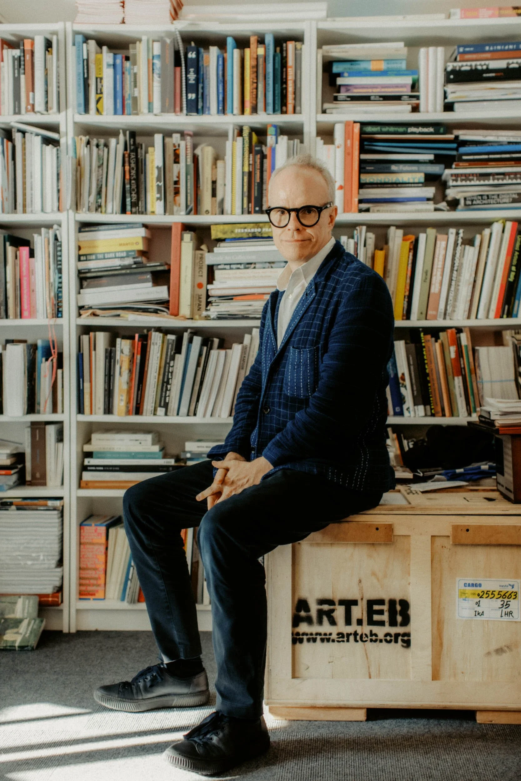 A man sitting on a wooden crate with graffiti and a website URL, in front of a large bookshelf filled with books. He is wearing a navy blue checkered suit, white shirt, black shoes, and round black glasses.