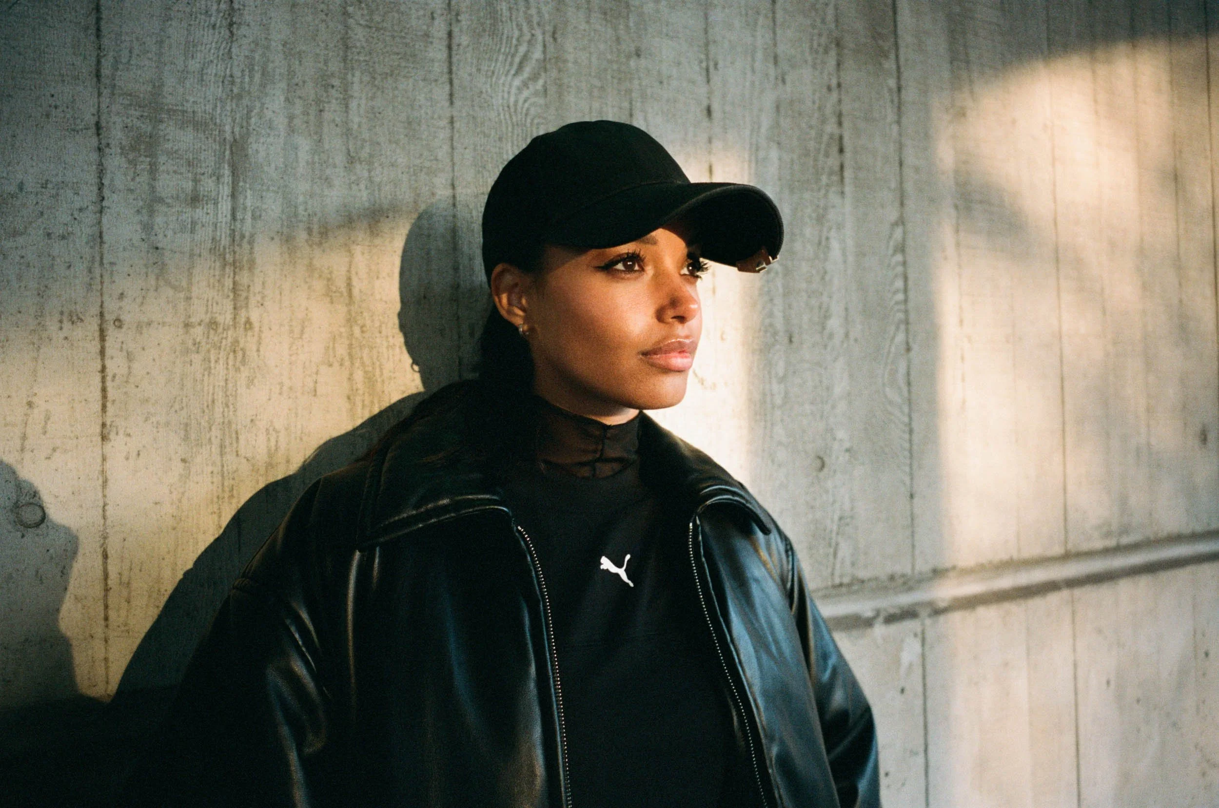 Linska with dark hair wearing a black cap, black leather jacket, and black shirt, standing against a concrete wall with her gaze directed to the right.
