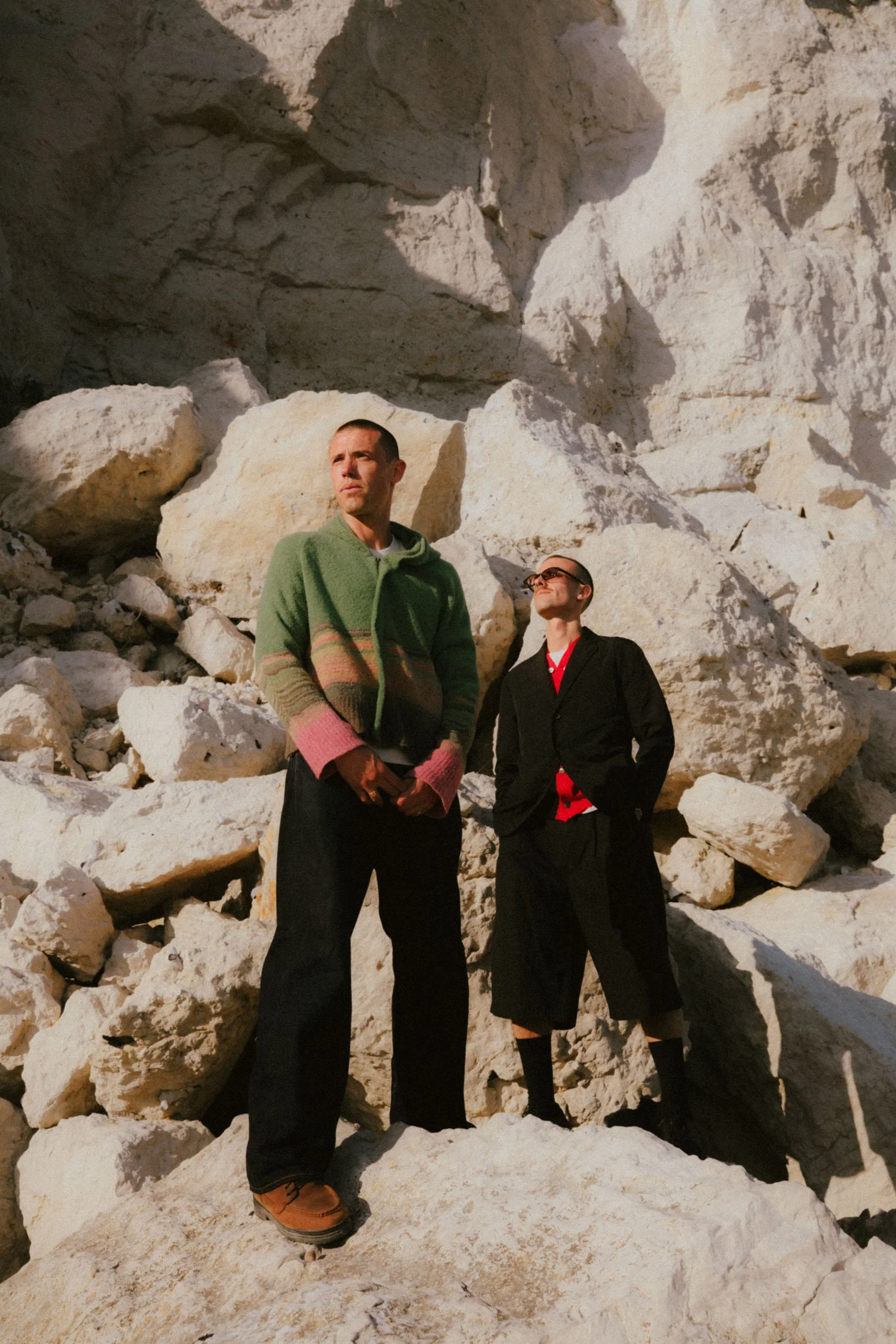 Two men standing among large rocks in a mountainous area with cliffs in the background.