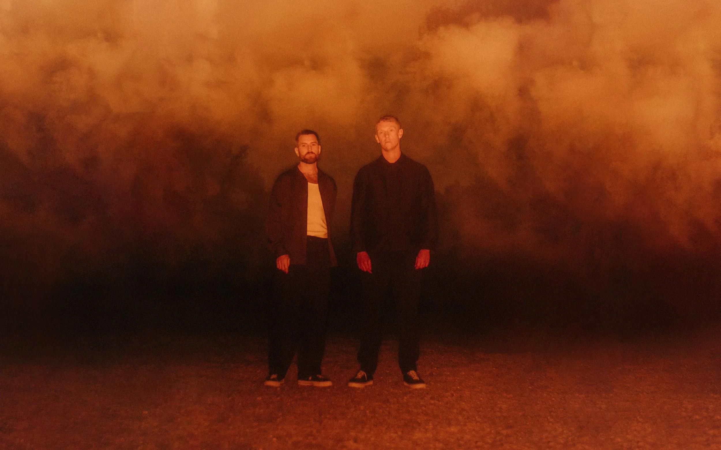 Two people standing outdoors at night with an orange, cloudy sky in the background.