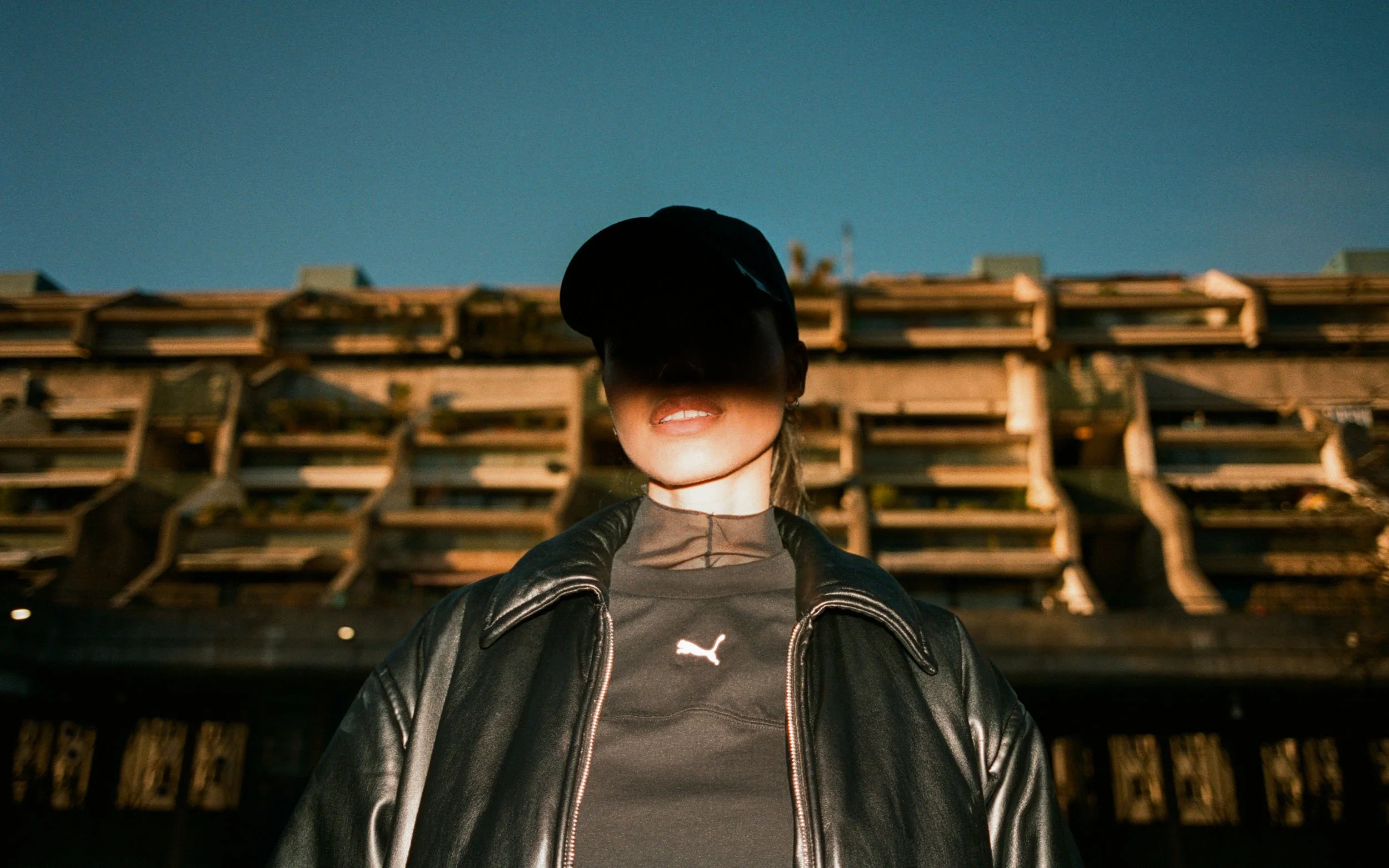 Person wearing a black cap, black leather jacket, and black Puma shirt standing outdoors in front of a wooden structure with a blue sky overhead.