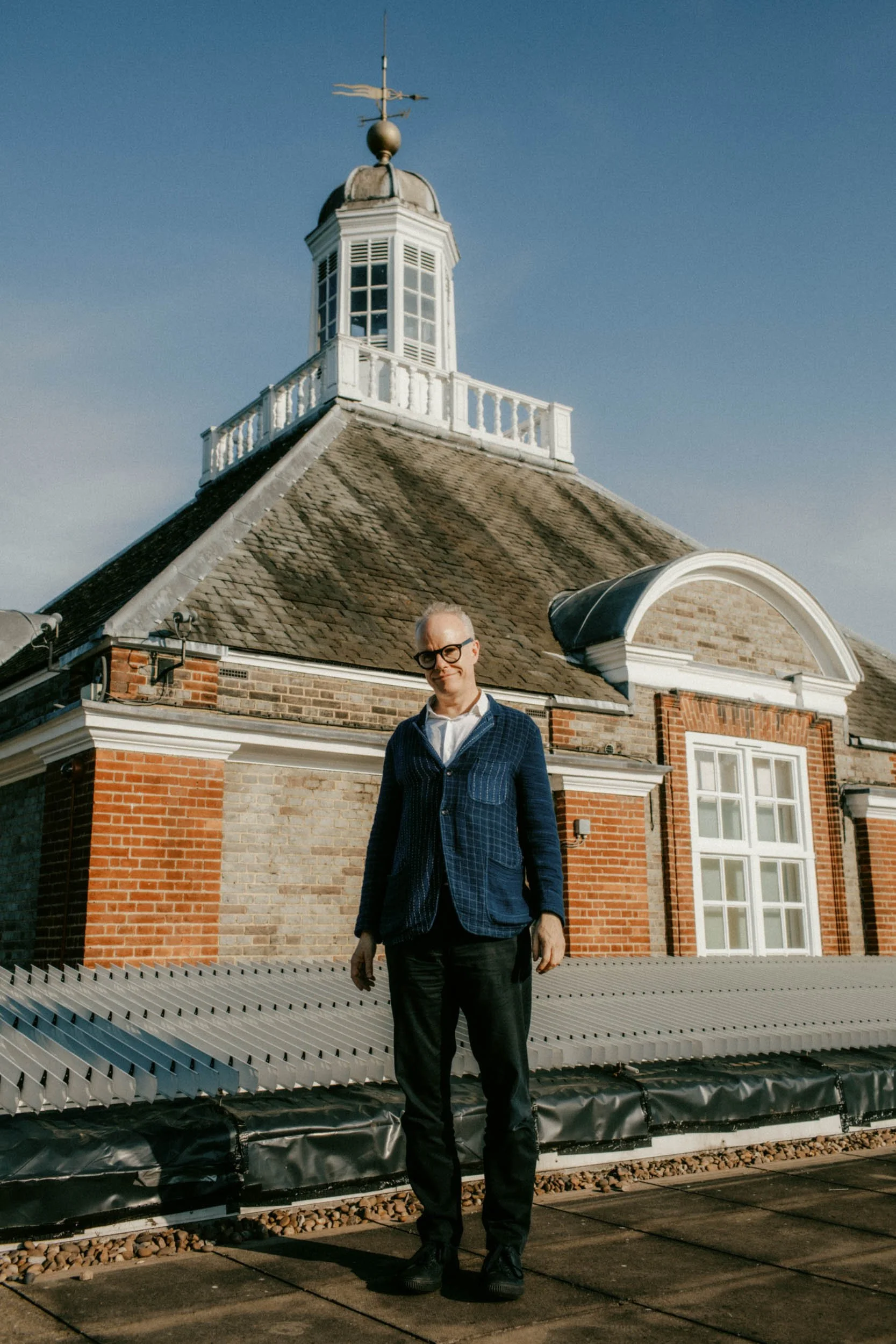 A man with glasses and gray hair stands on a building rooftop with a solar panel array in the foreground, and a brick building with a clock tower and cupola with weather vane under a clear blue sky.
