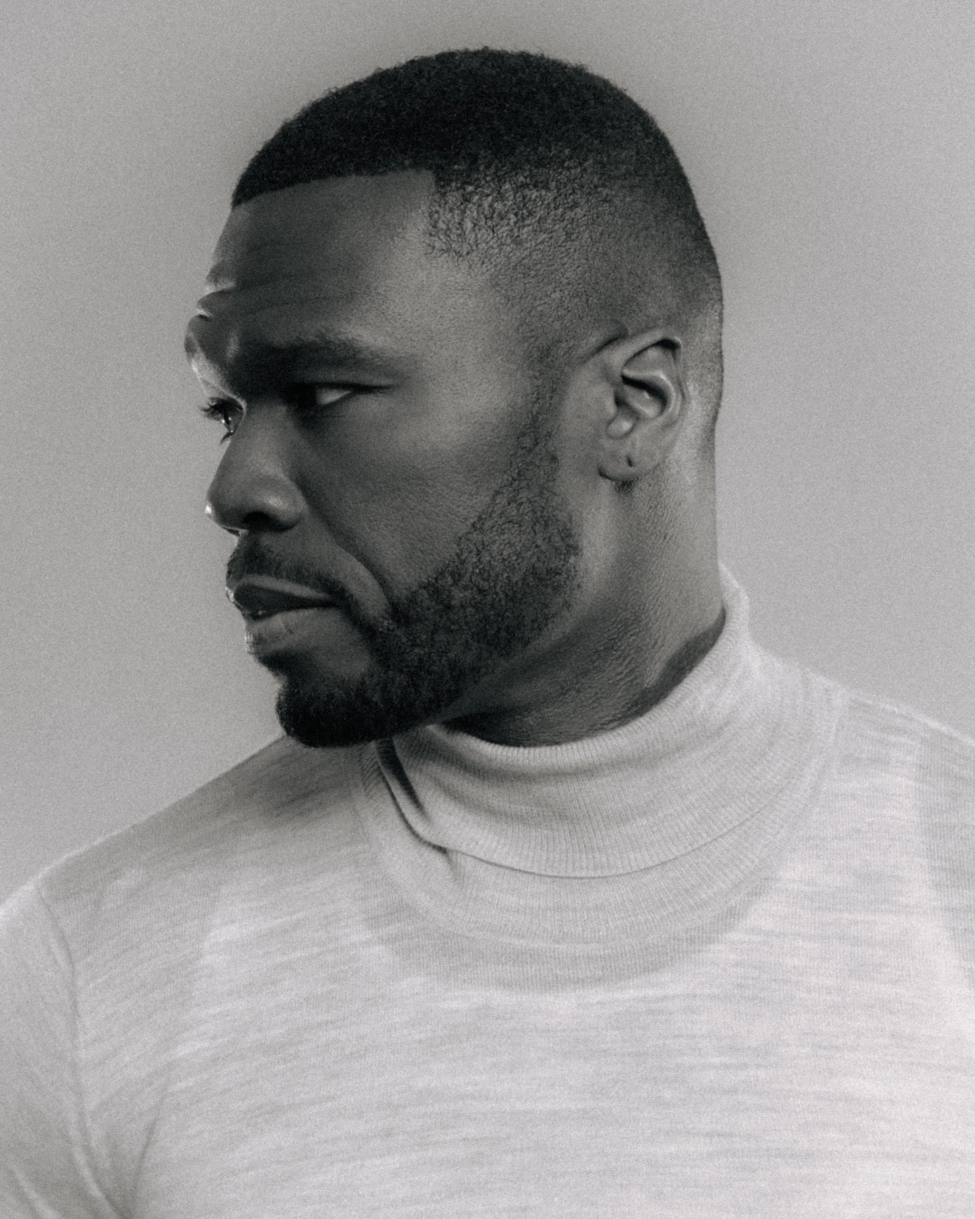Close-up black and white photo of a man's face in profile, showing hairstyle, ear, and part of a T-shirt, with a neutral background.