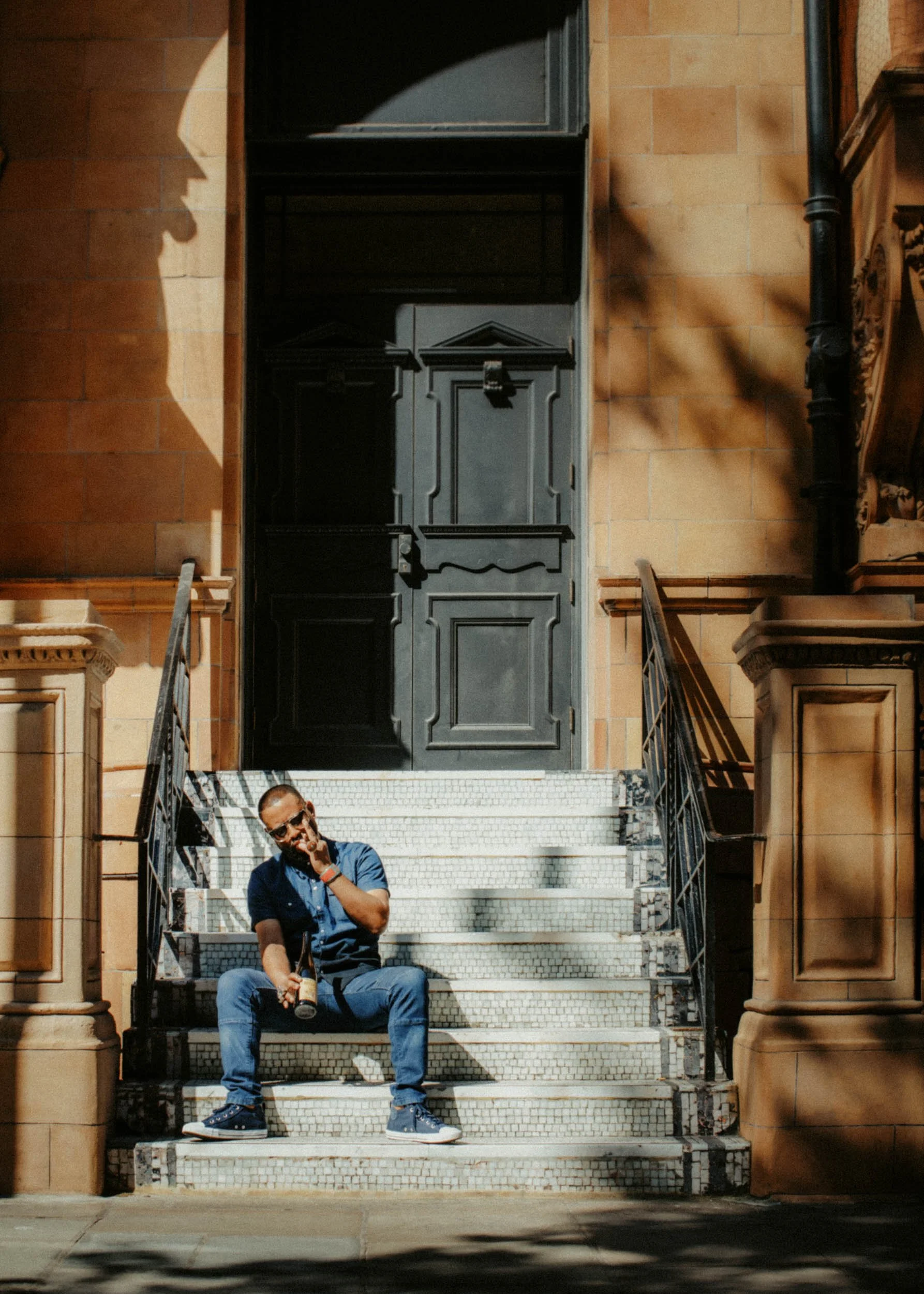 A man sitting on the stairs outside a building, holding a beer bottle, wearing sunglasses, a blue shirt, jeans, and sneakers, with shadows cast on the building's exterior and stairs.