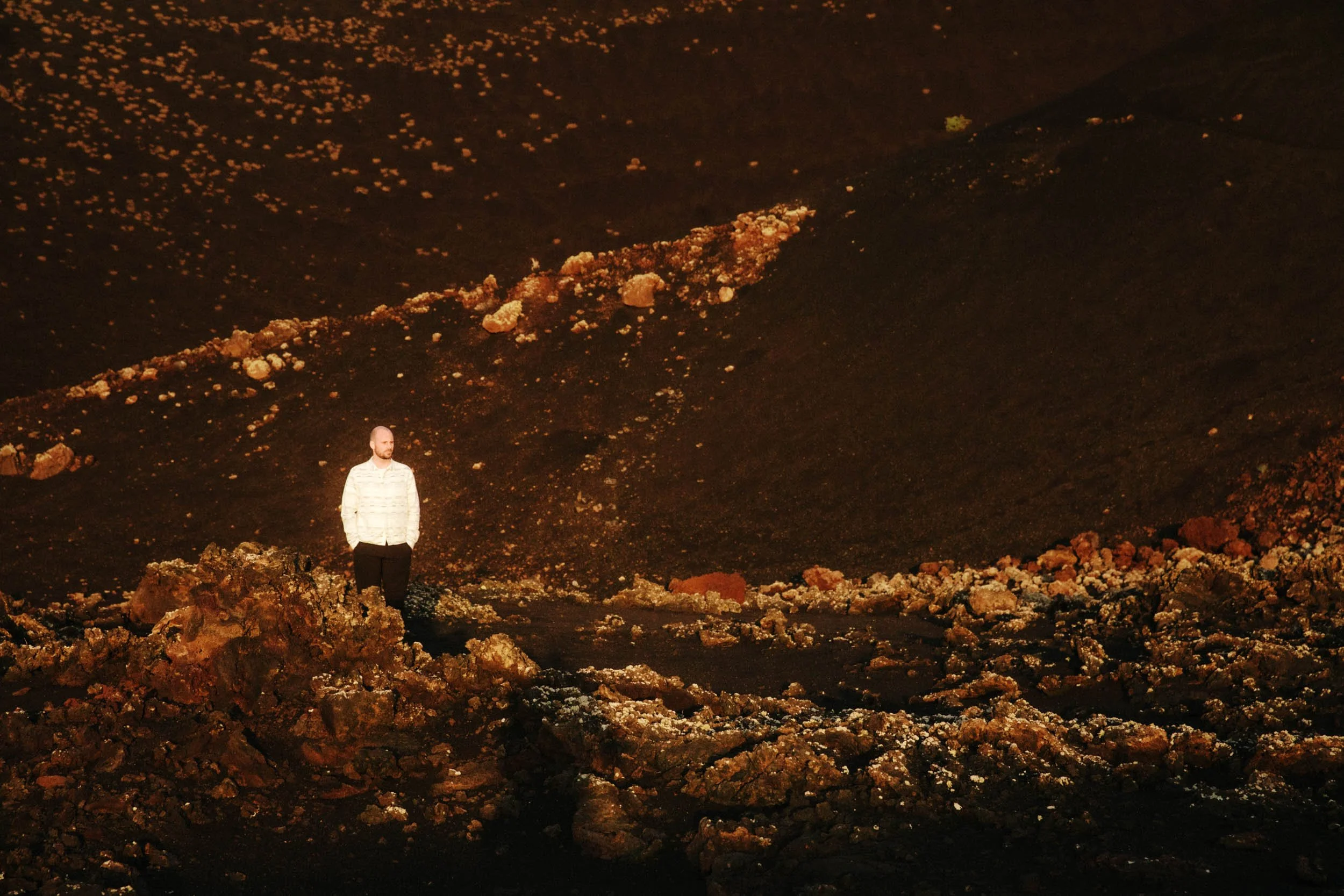 A man with a beard and striped shirt standing on a rocky landscape with dark volcanic terrain and scattered rocks, possibly on a lava field or similar environment.