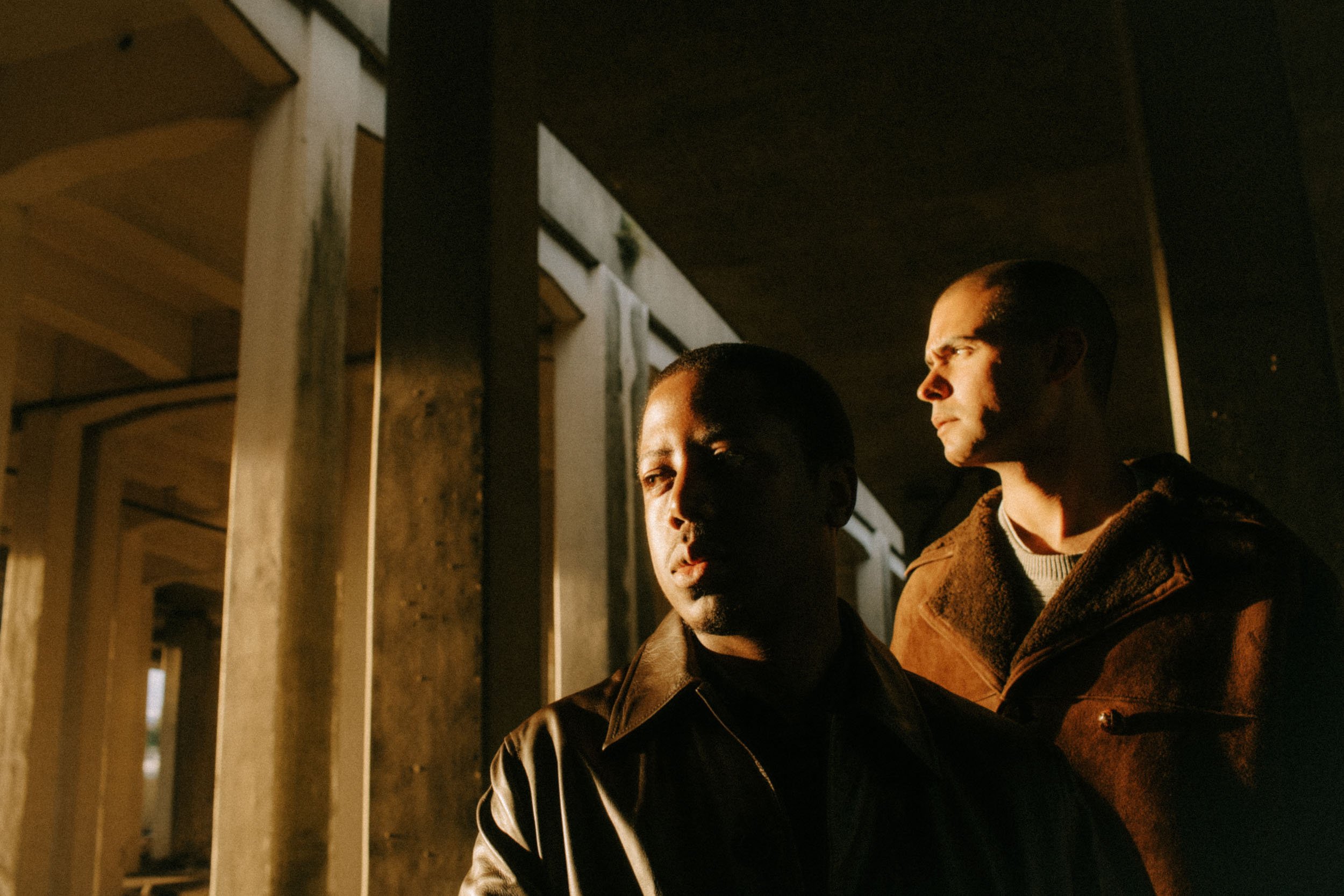 Two men with closely shaved heads, one with darker skin and the other with lighter skin, standing outdoors at night under a bridge or overpass with concrete pillars. The scene is dimly lit.