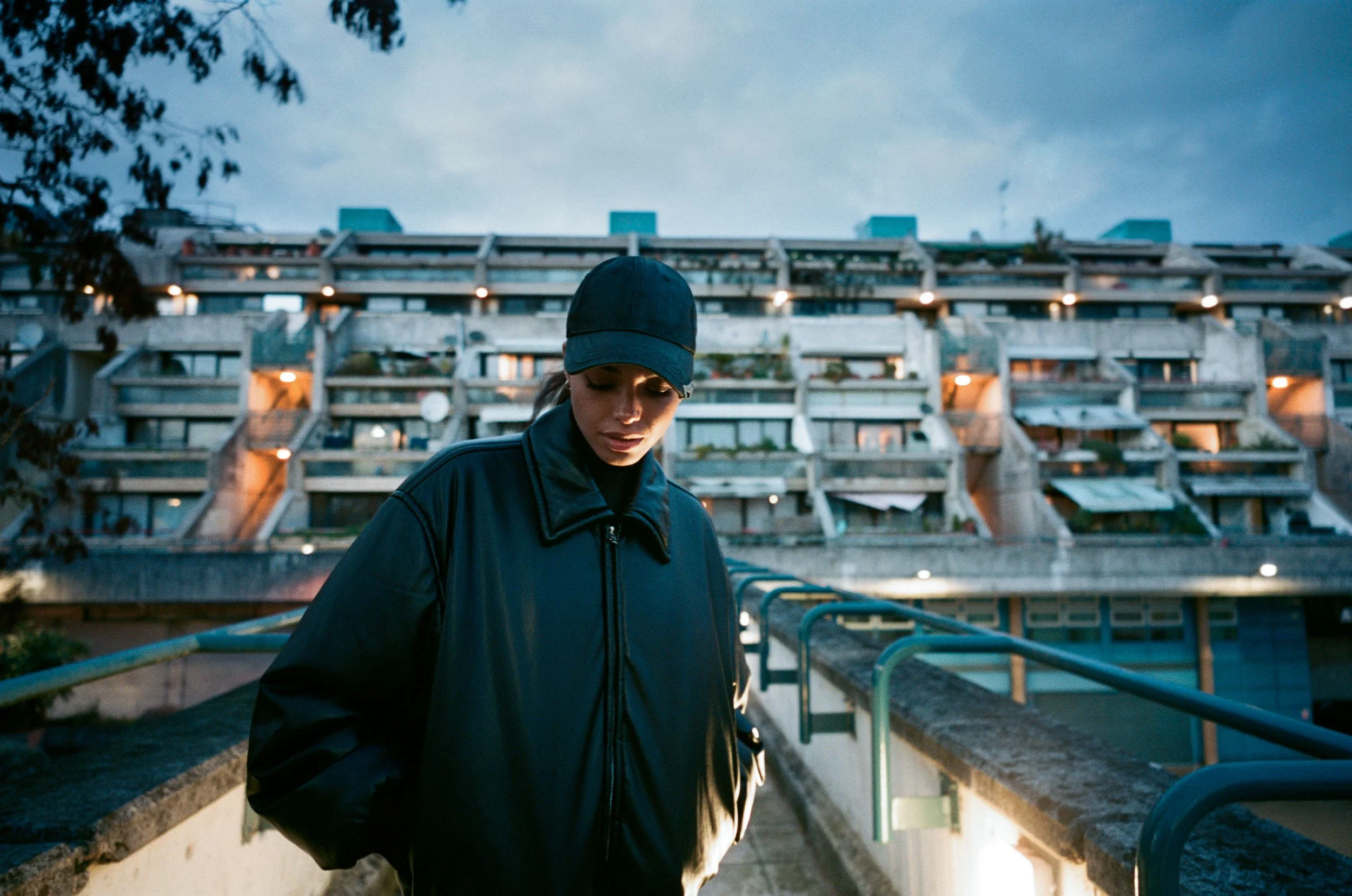 Linska wearing a black cap and jacket stands outdoors beneath a cloudy evening sky in front of a multi-level parking structure with blue accents and lights.