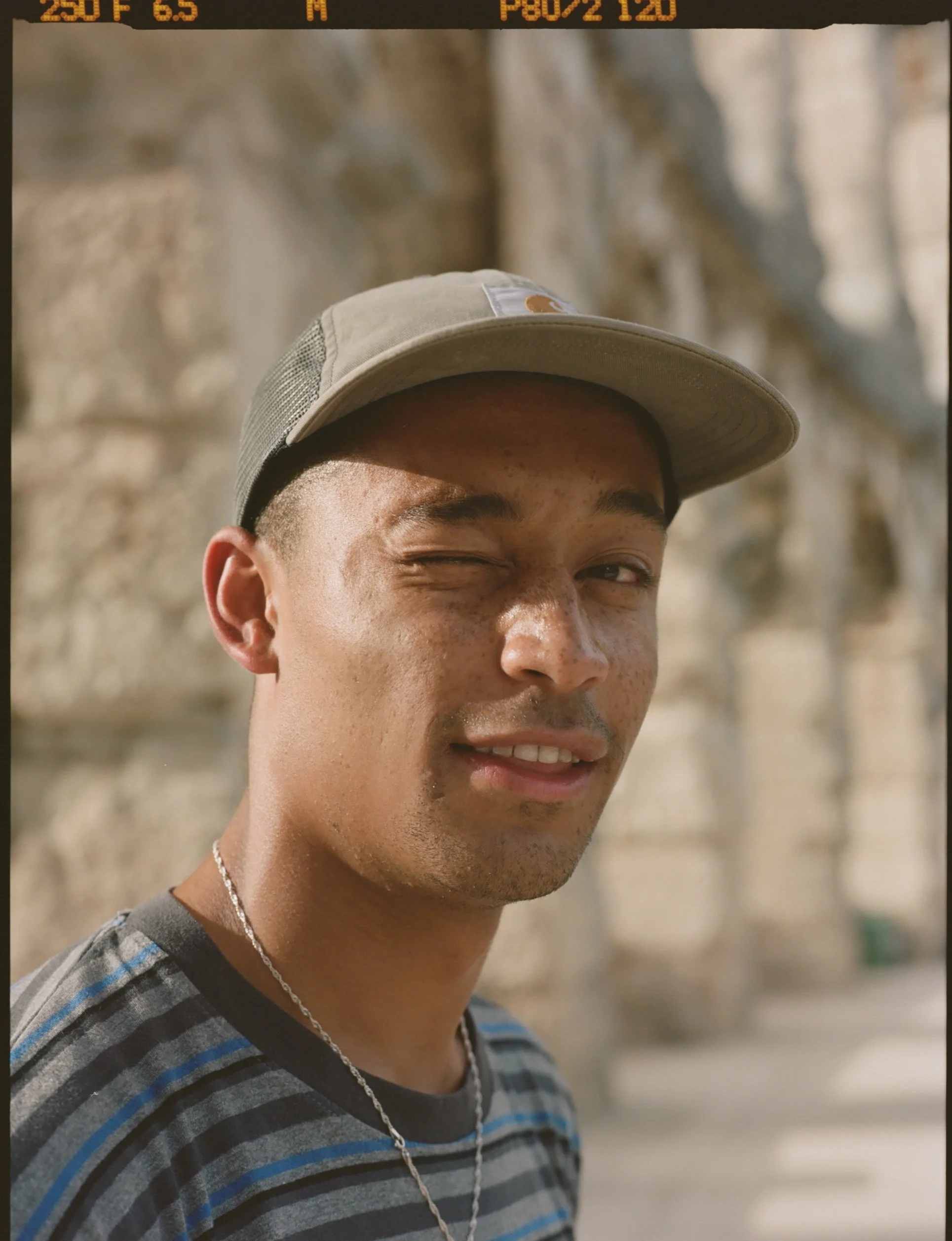 A young man wearing a beige baseball cap with a brown logo, a striped gray and blue t-shirt, and a silver chain necklace, smiling and winking at the camera. The background is blurry and appears to be an outdoor setting.
