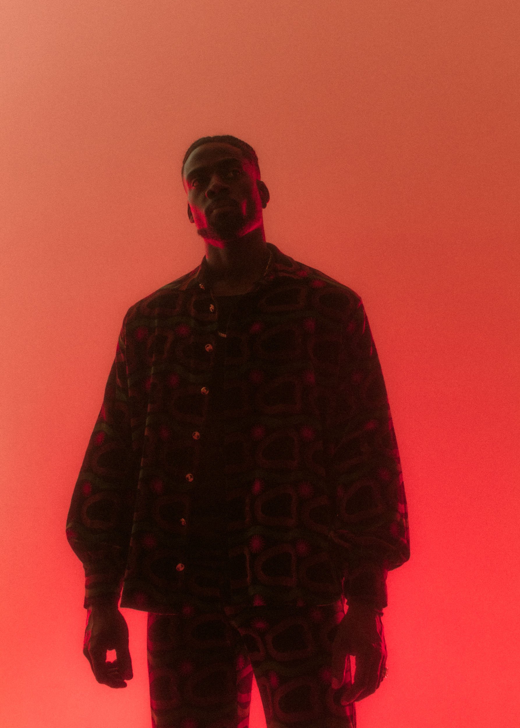 A young man with braided hair and a beard stands against a red-lit wall, wearing a patterned jacket and pants with a similar design, looking directly at the camera.