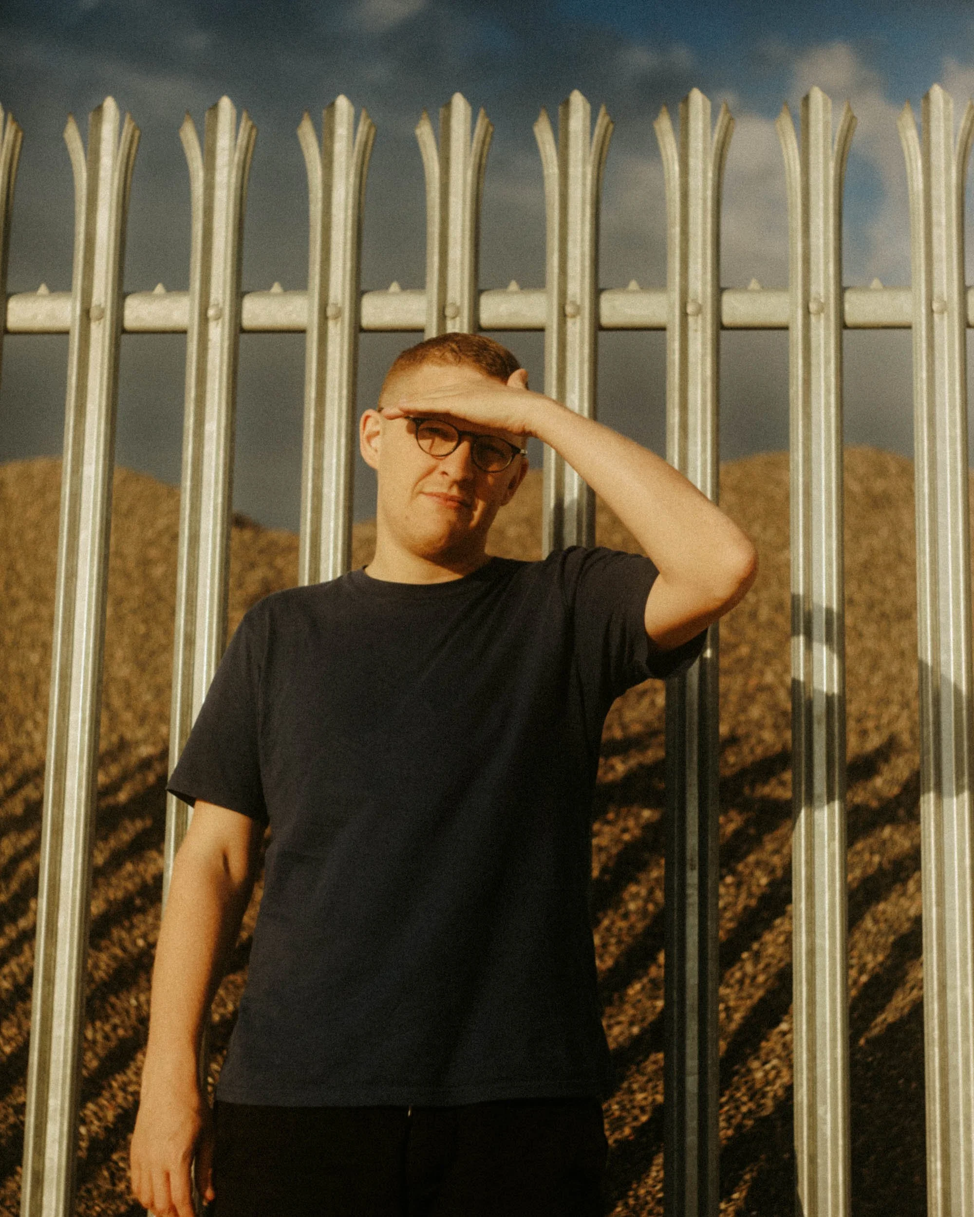 A young man with glasses, short hair, wearing a black t-shirt, shielding his eyes with his right hand, standing behind a metal fence outdoors with mountains and a cloudy sky in the background.
