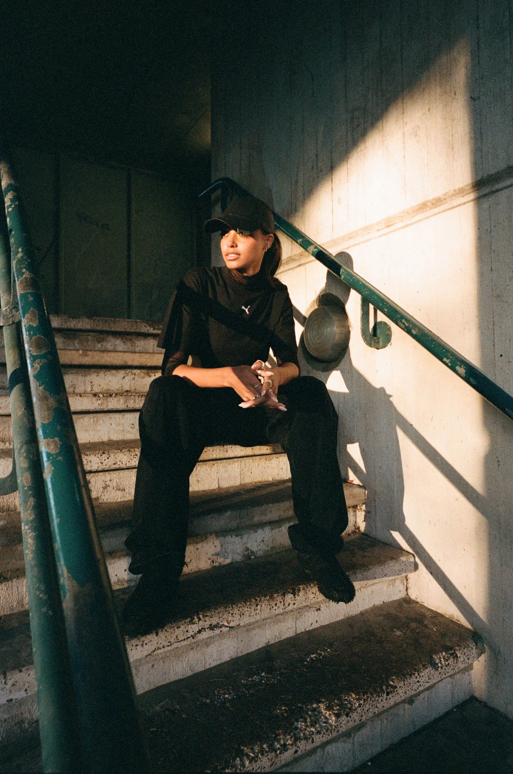 A woman sitting on concrete stairs, wearing a black outfit and baseball cap, with her hands clasped and looking to the side, illuminated by sunlight with a concrete wall behind her.