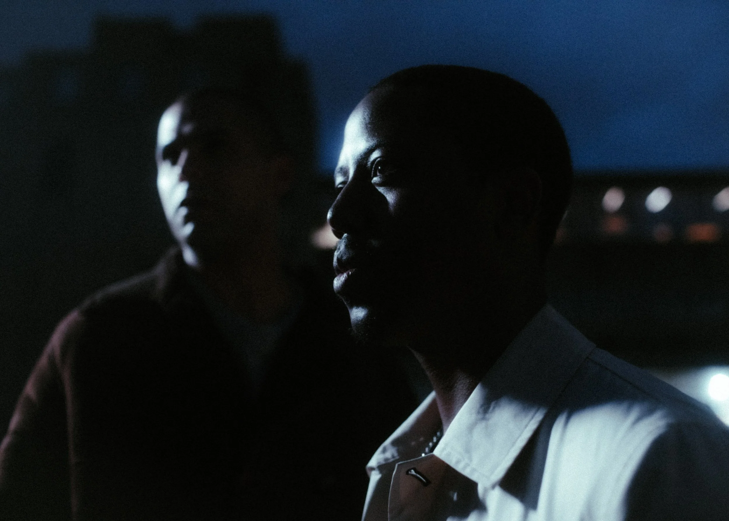 A close-up of a man and woman in a dimly lit setting, with their faces partially illuminated, showing the woman in focus on her profile, and the man slightly out of focus in the background.