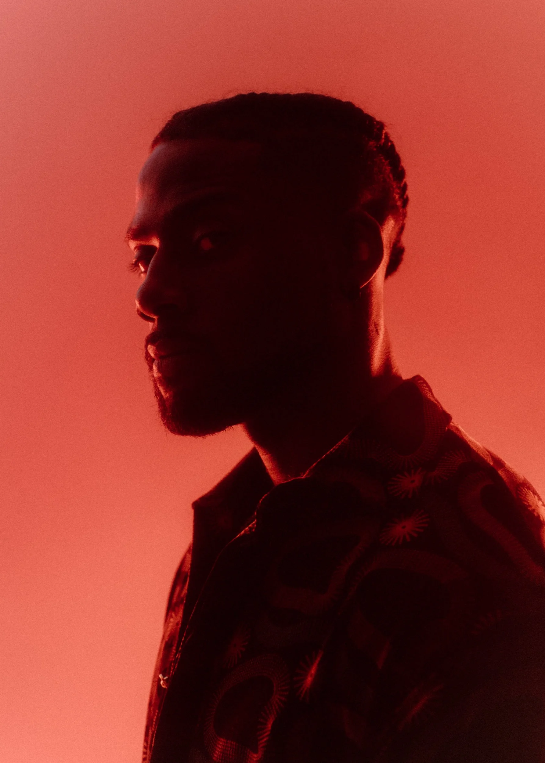 Side profile of a man with short curly hair illuminated by red lighting, wearing a shirt with a floral pattern.