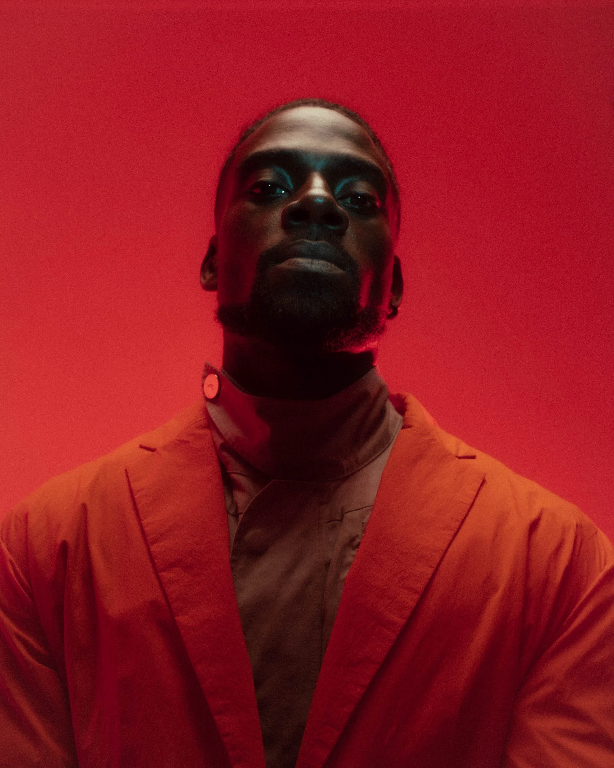 A person with dark skin, short hair, and a beard wearing an orange jacket over a patterned shirt, standing against a red background.