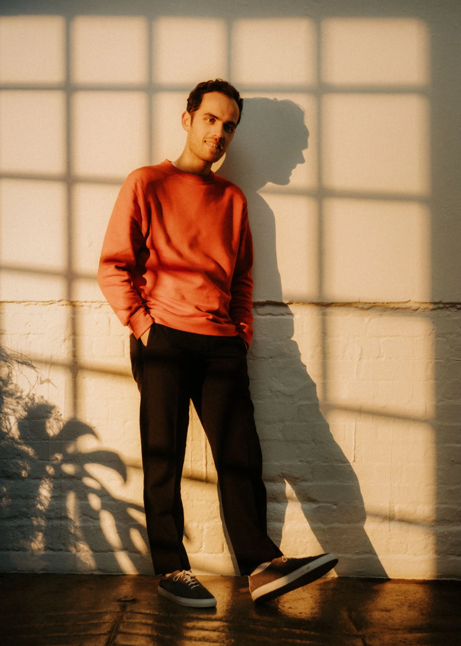 A young man with dark hair and a slight smile, wearing a reddish long-sleeve shirt and black pants, stands against a white brick wall. The setting appears to be indoors with warm sunlight casting a shadow of his profile on the wall, creating grid-lik
