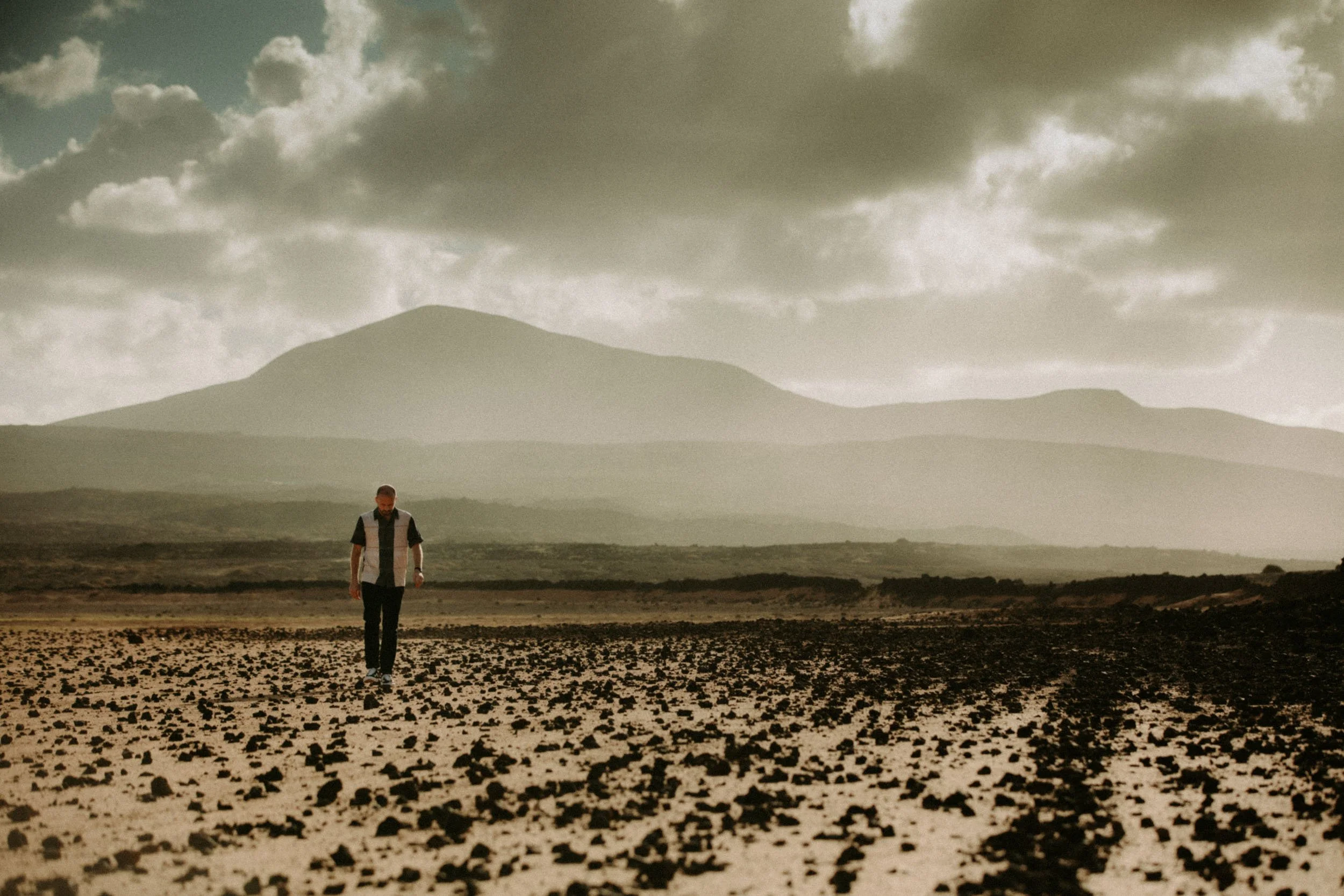 A man walking alone in a dry, rocky field with mountains and cloudy sky in the background.