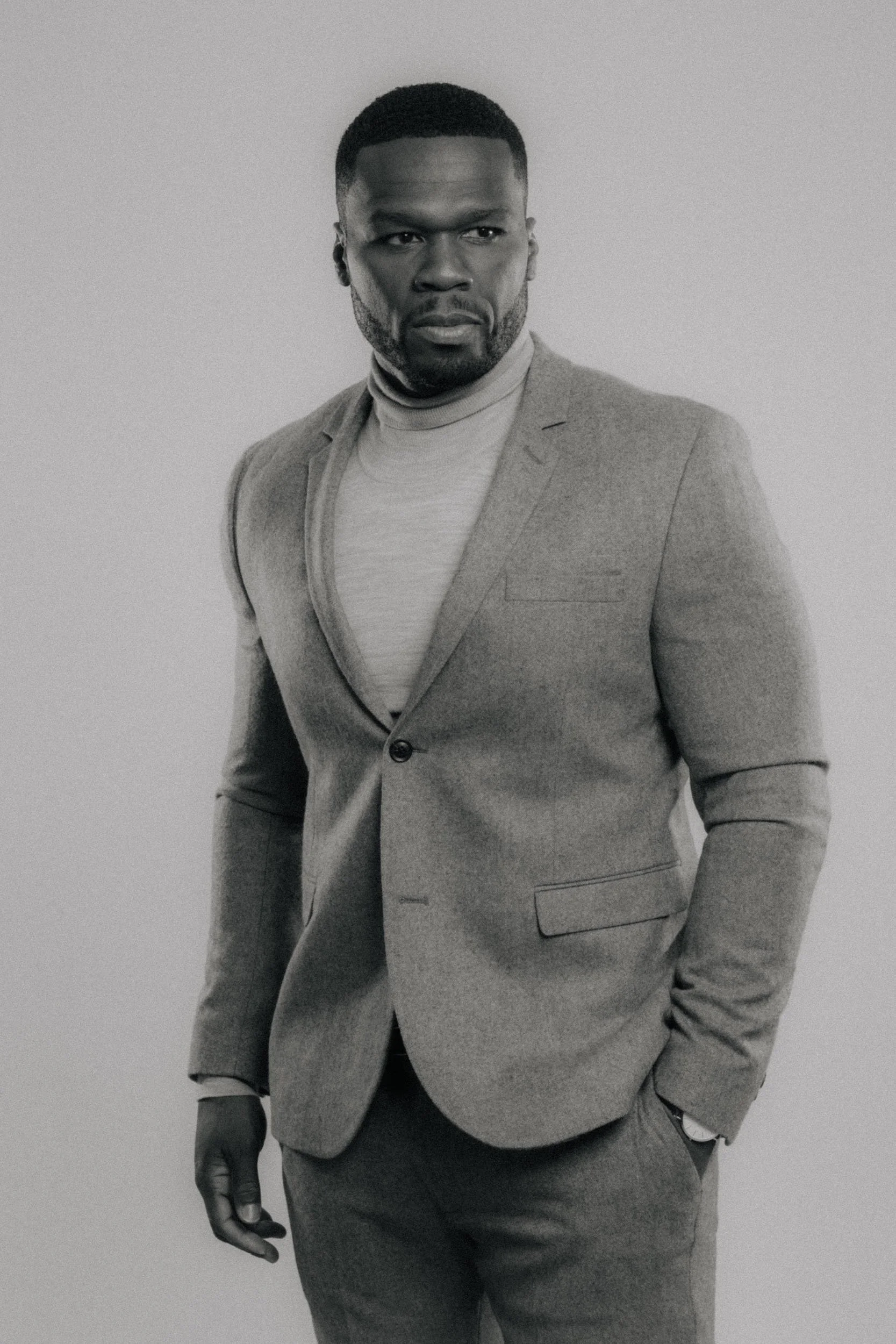 Black and white photo of a man in a suit with a turtleneck, standing against a plain background, with a serious expression and one hand in his pocket.