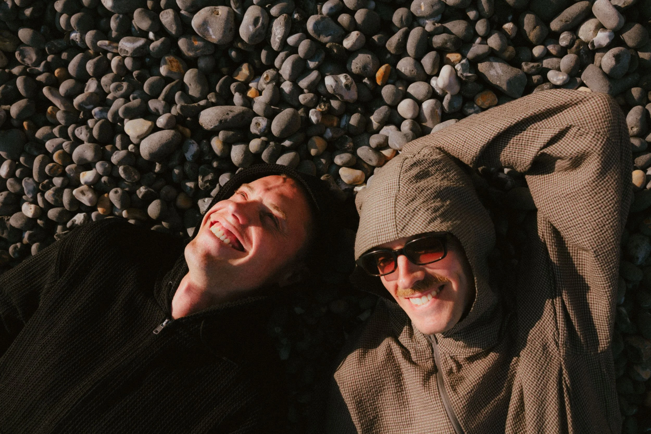 Two people lying on a bed of small rocks, smiling and taking a selfie.