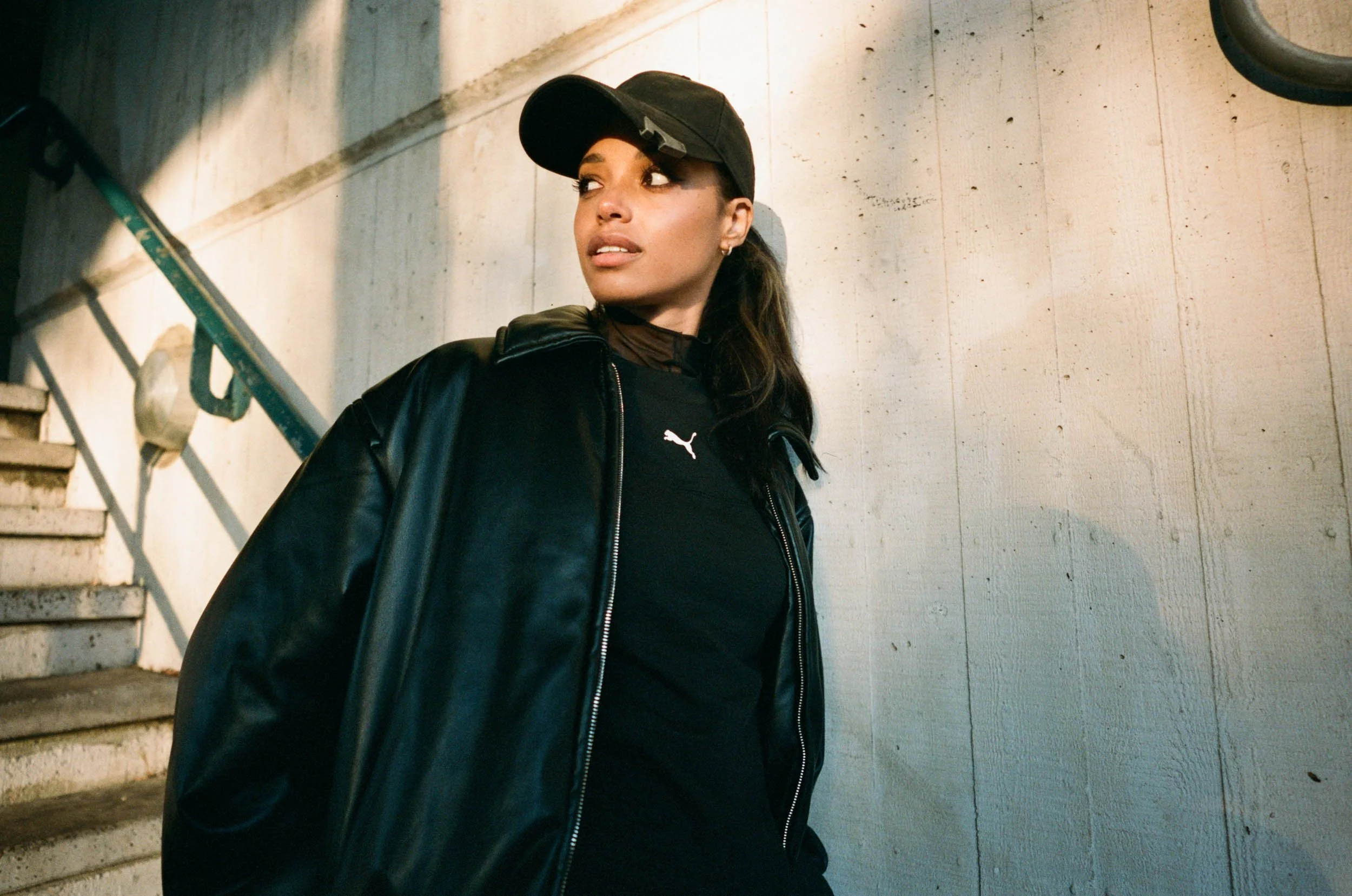 A woman wearing a black jacket, black cap, and black Puma shirt, standing against a concrete wall near a staircase with metal railing.