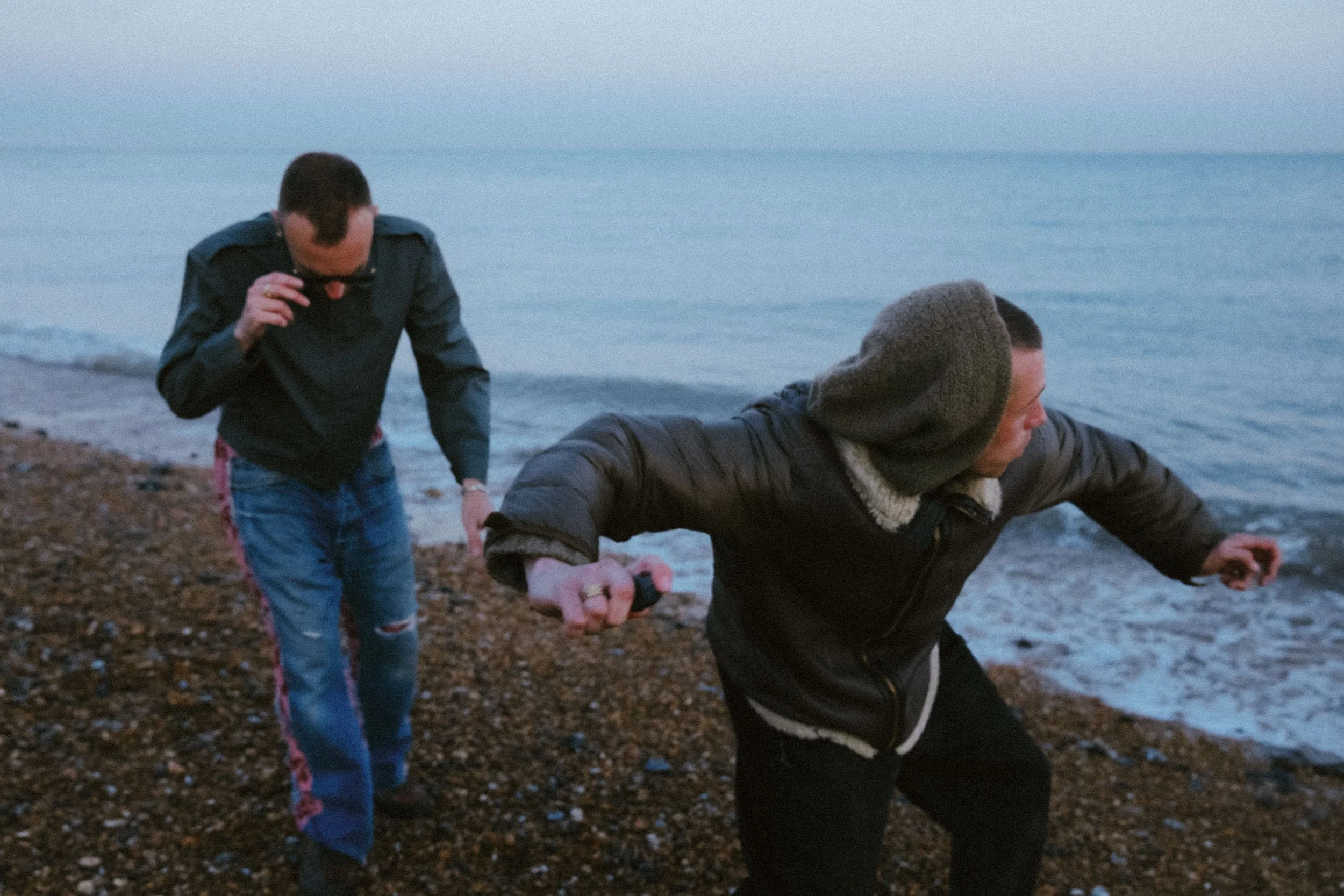 Two men on a pebble beach near the water during dusk, one wearing a hoodie and the other a jacket, holding hands.